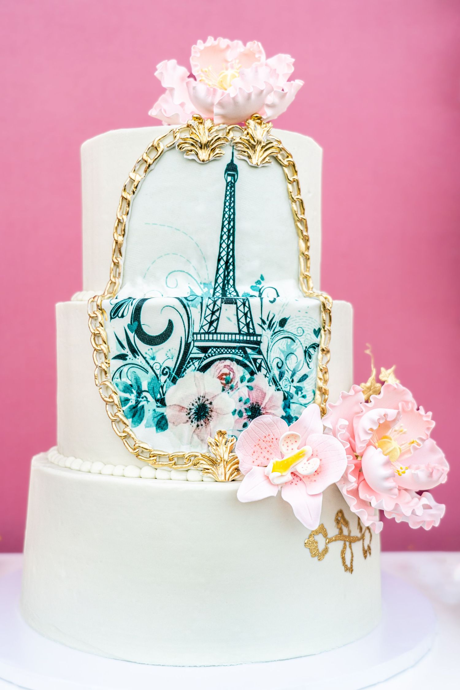 Elegant wedding cake with a hand-painted Eiffel Tower design in turquoise and gold trim against a pink background.