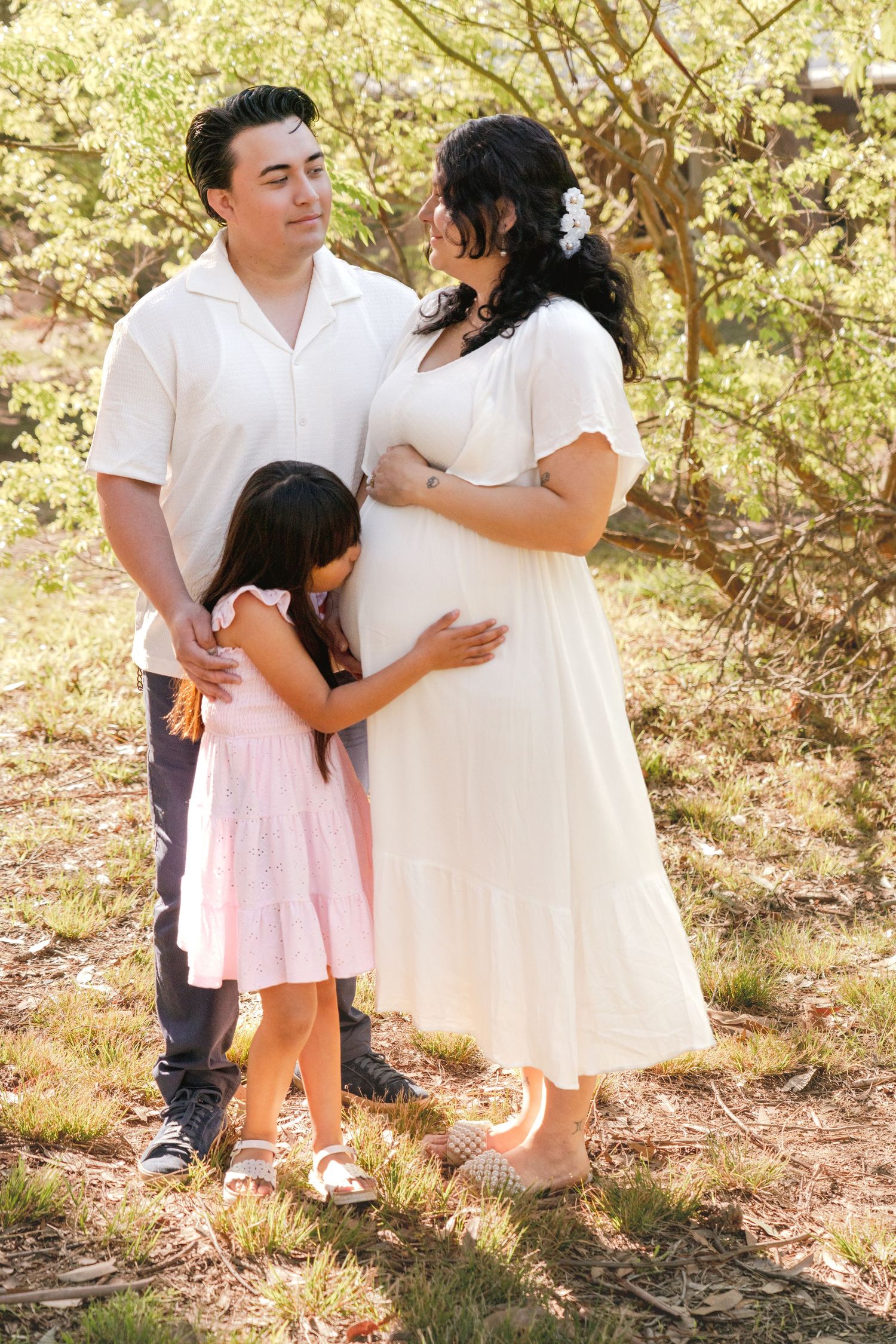 Family wearing white outfits poses together in a sun-dappled wooded setting.
