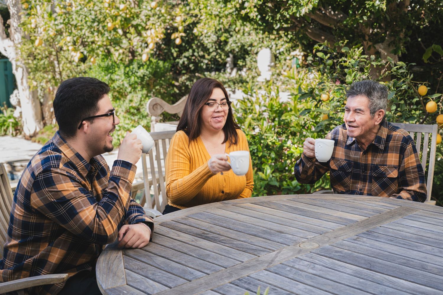 Family sharing laughs while sitting at outdoor wooden table in garden setting.