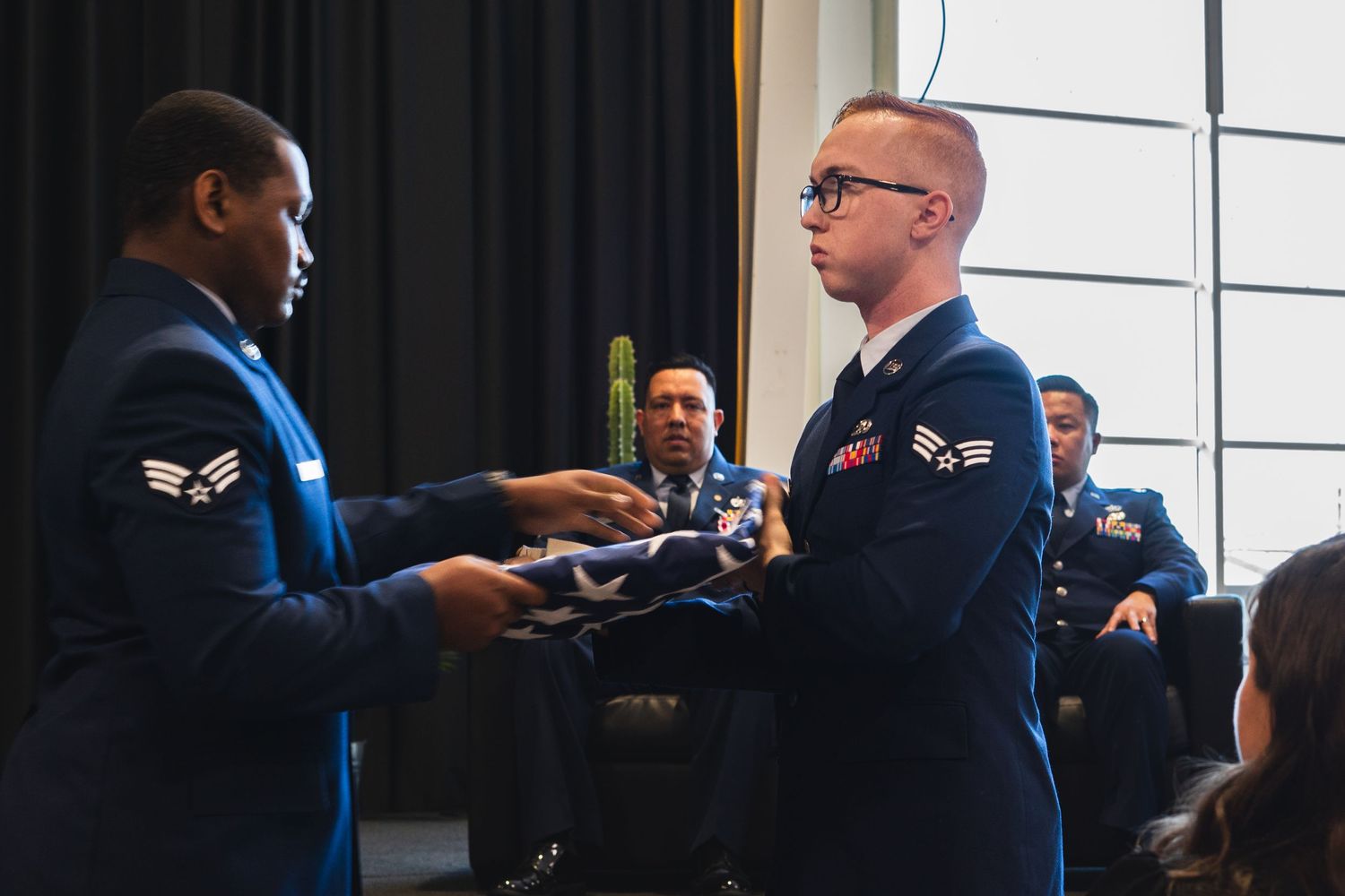 Two Air Force airmen in uniform exchange a folded flag during a military ceremony indoors.