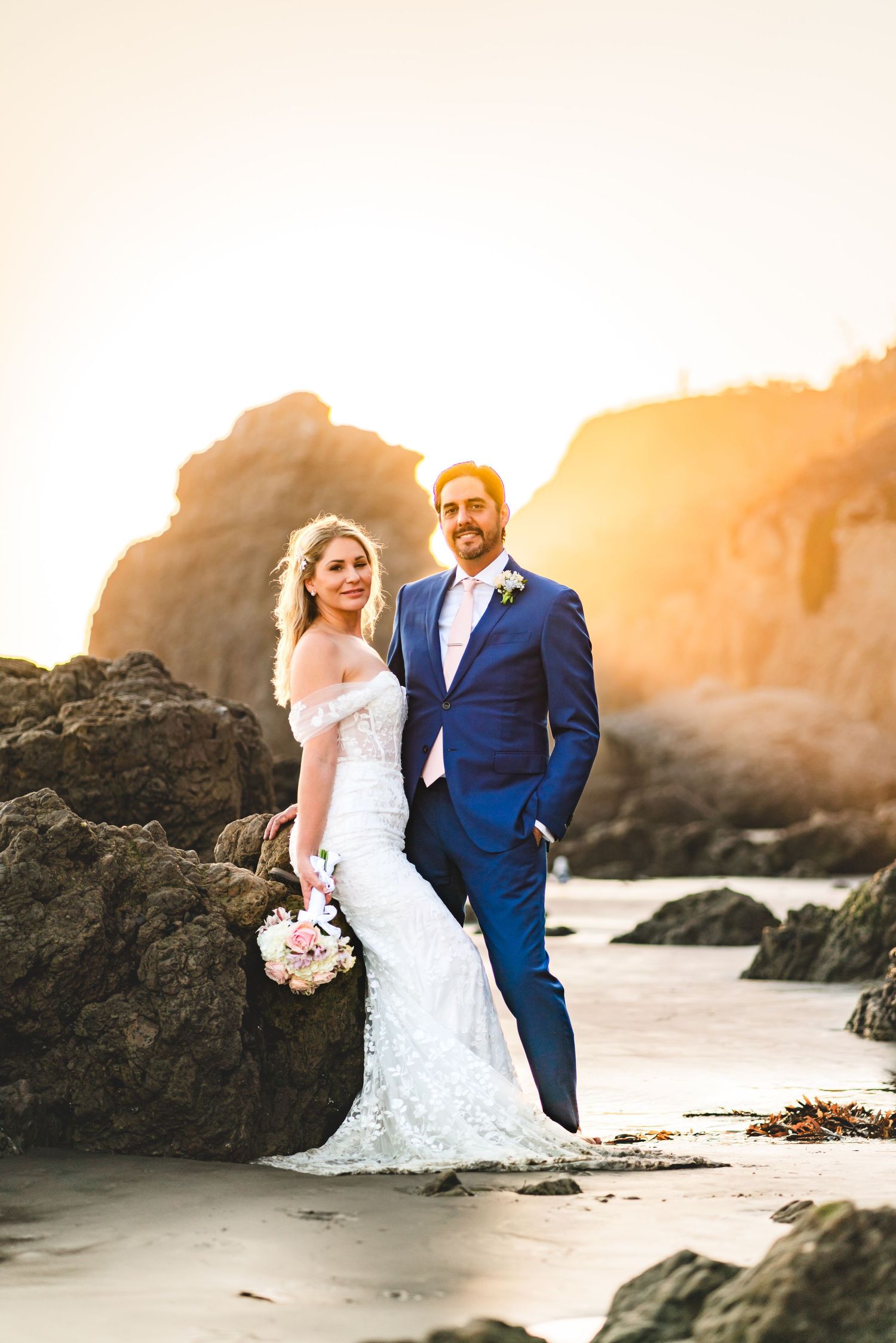 A romantic beach wedding photoshoot at sunset with a couple posing among coastal rocks in formal attire.