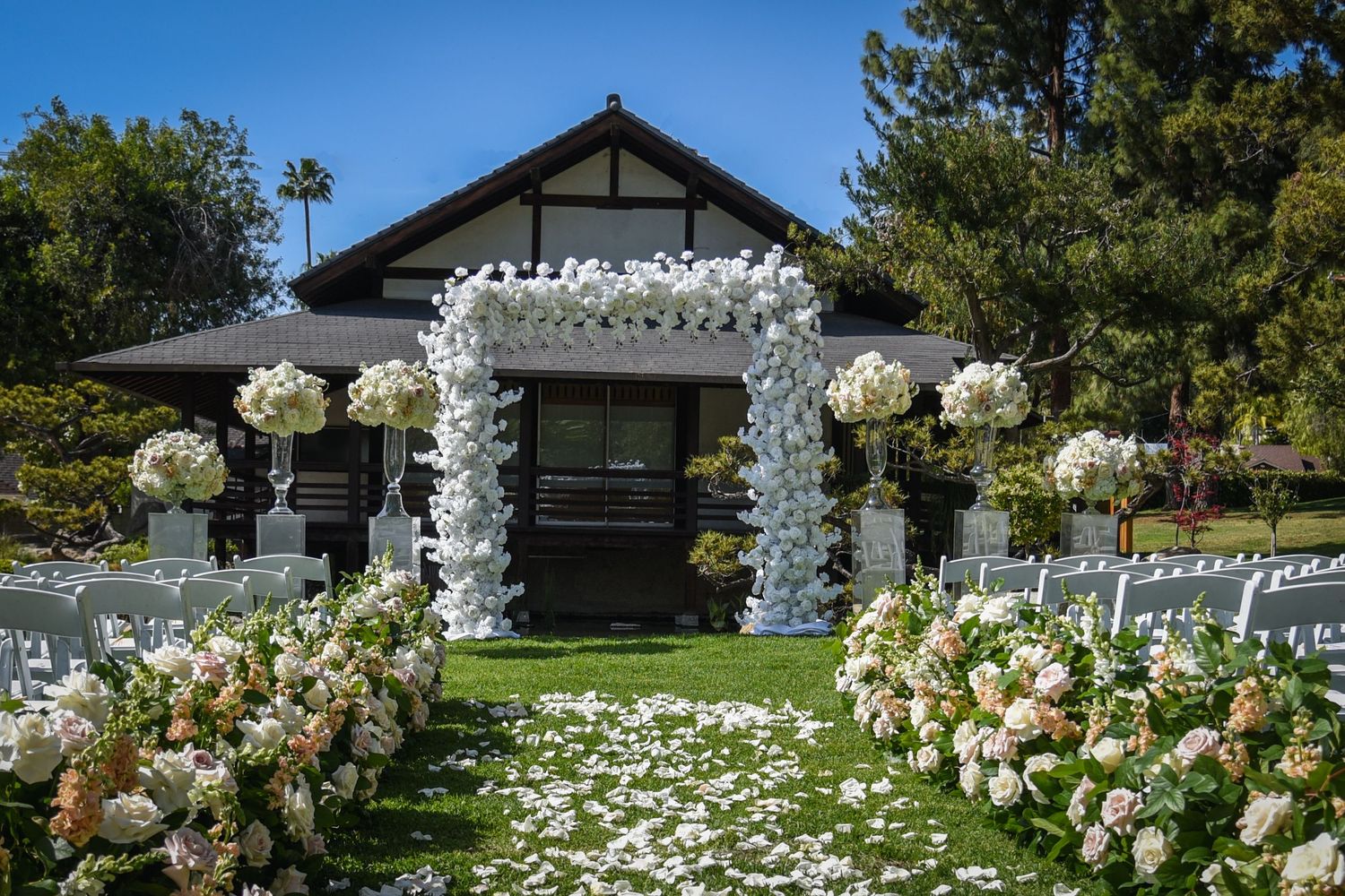 Outdoor wedding ceremony setup with white floral arch and garden chairs against a blue sky.