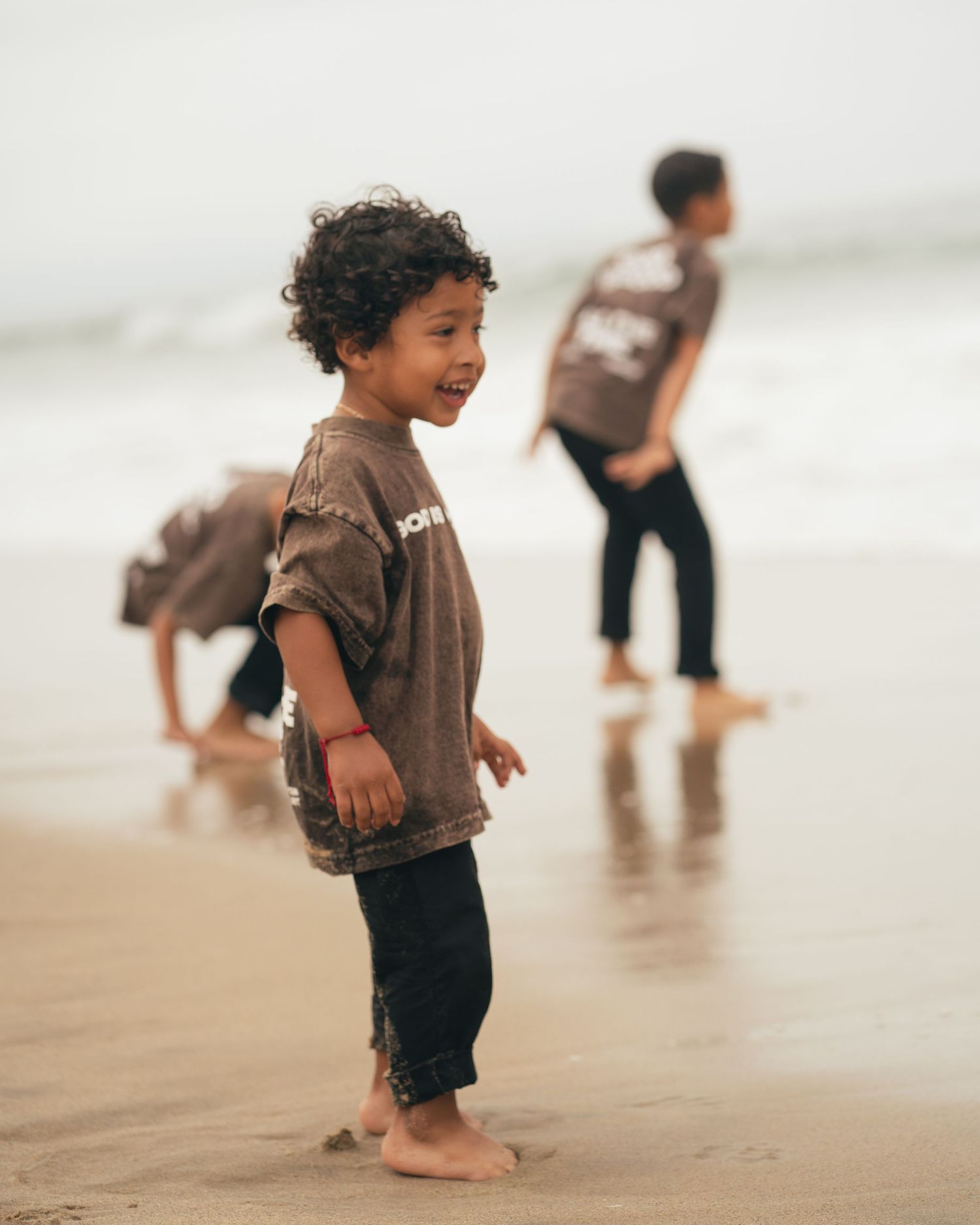 A joyful young child smiles and plays barefoot on a sandy beach with other children playing in the background.