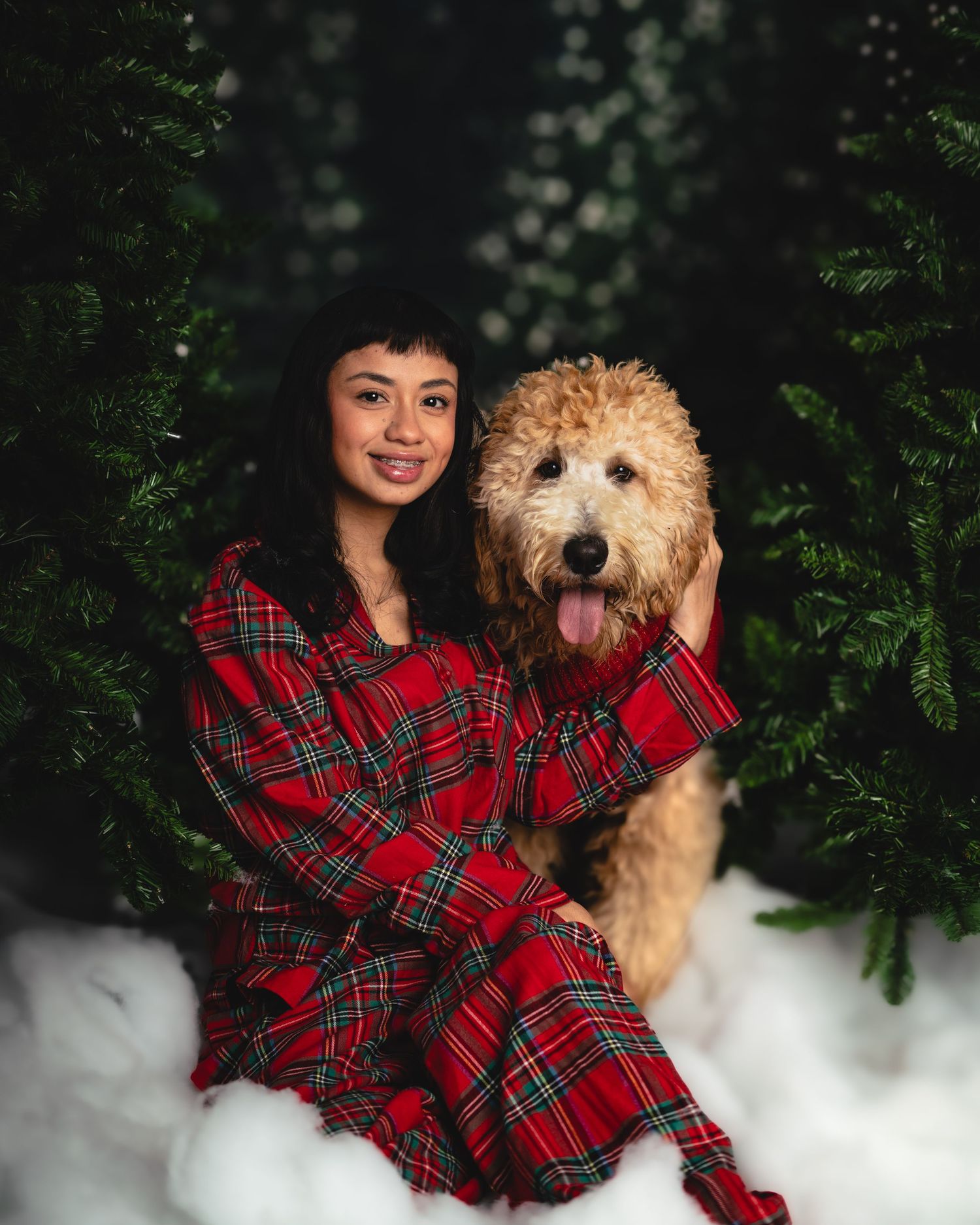 A person in a red plaid shirt poses with a fluffy goldendoodle dog against a dark green foliage background.
