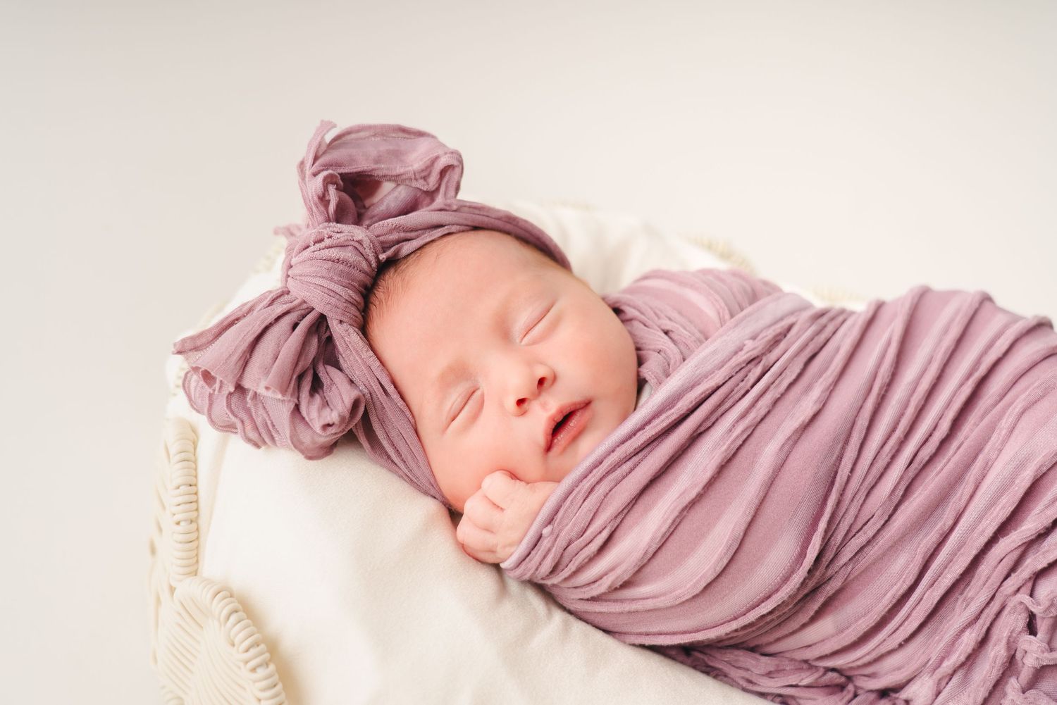 Baby sleeping soundly wrapped in flowing purple fabric against white backdrop.