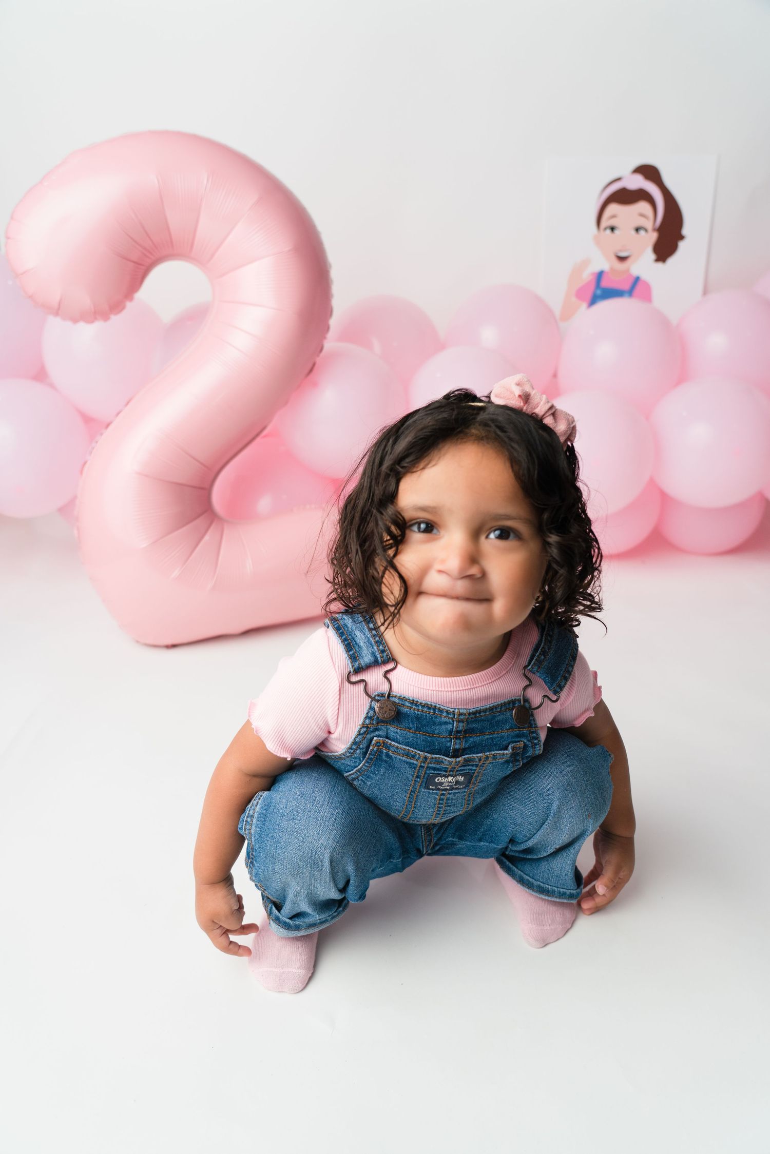A small child in denim overalls plays on the floor with pink heart-shaped pillows in a white studio setting.