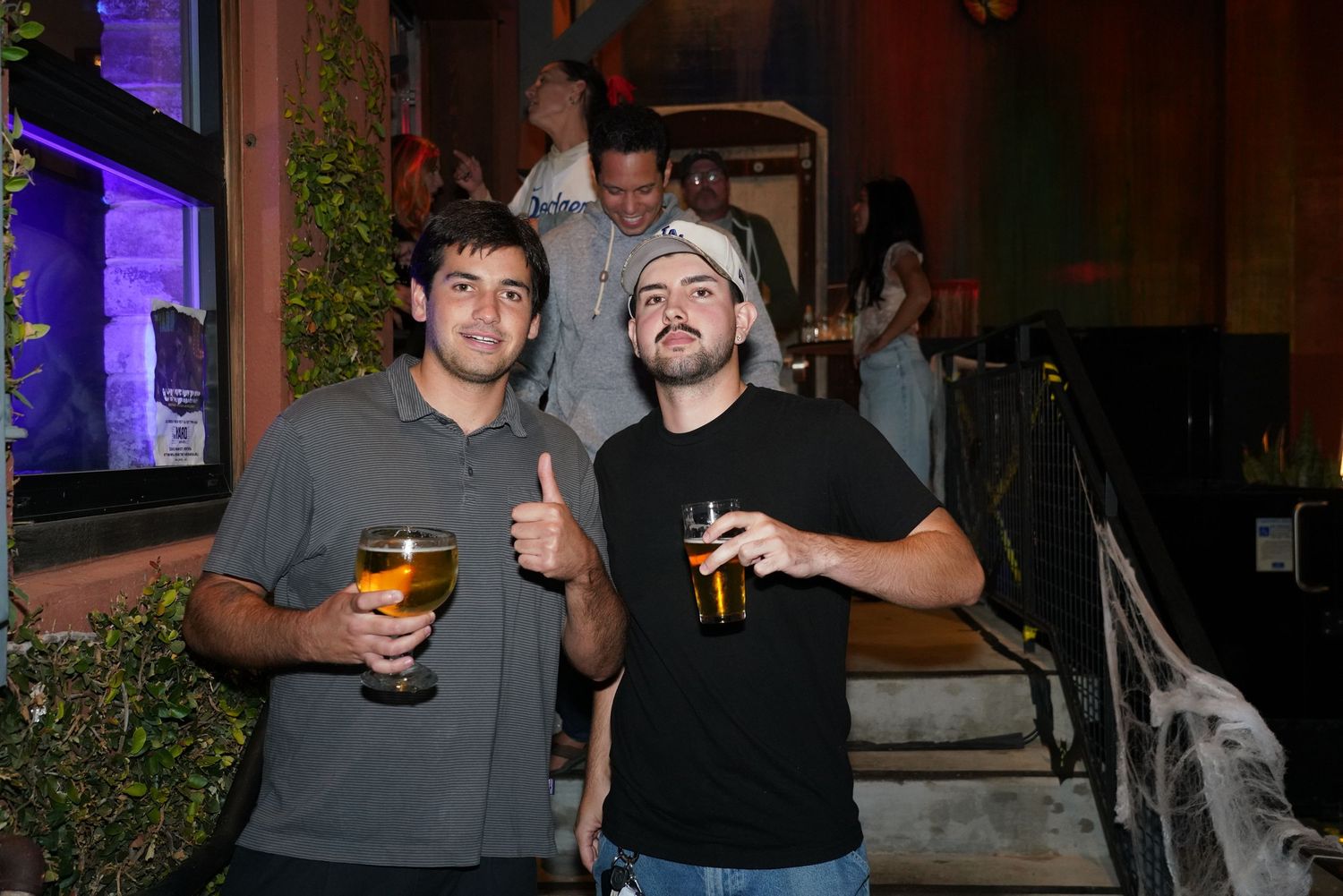 Two young men smiling and holding drinks at an outdoor bar or patio venue at night.
