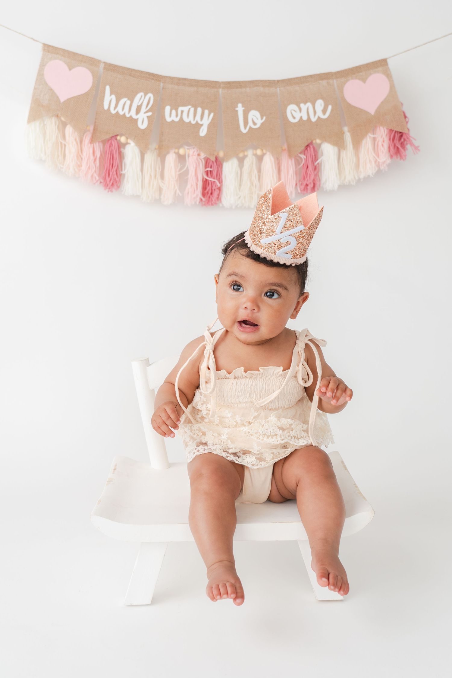 Baby in lace romper sitting beneath 'half way to one' banner with pink and white decorative garland.