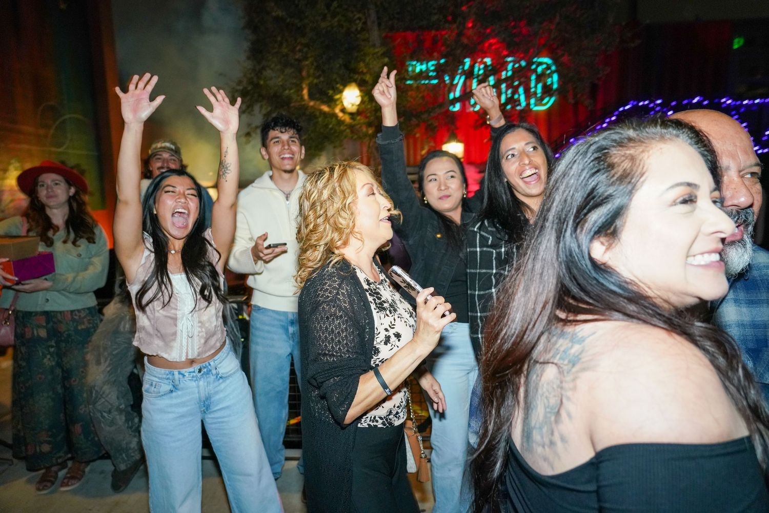 Excited crowd of people dancing and celebrating with raised hands at an outdoor nighttime event.