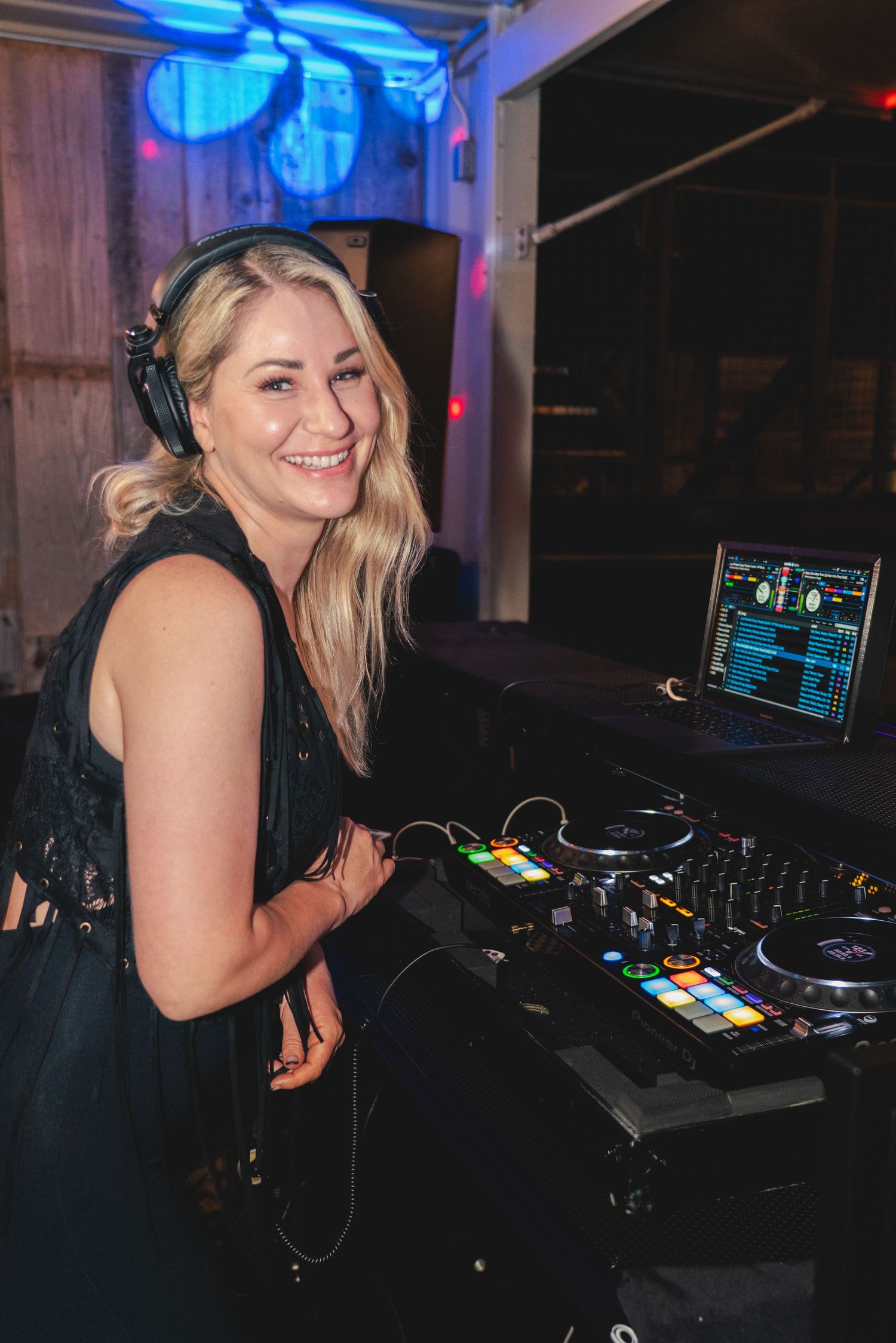 Smiling female DJ with headphones mixing music on a DJ controller with a laptop at a nightclub event.