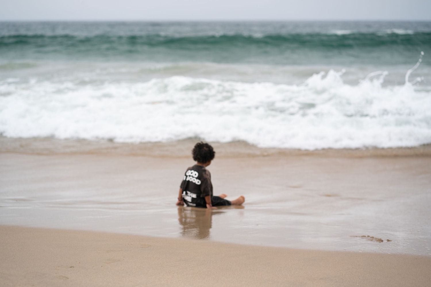 A young child sits alone on a sandy beach, watching ocean waves crash on the shore on a cloudy day.