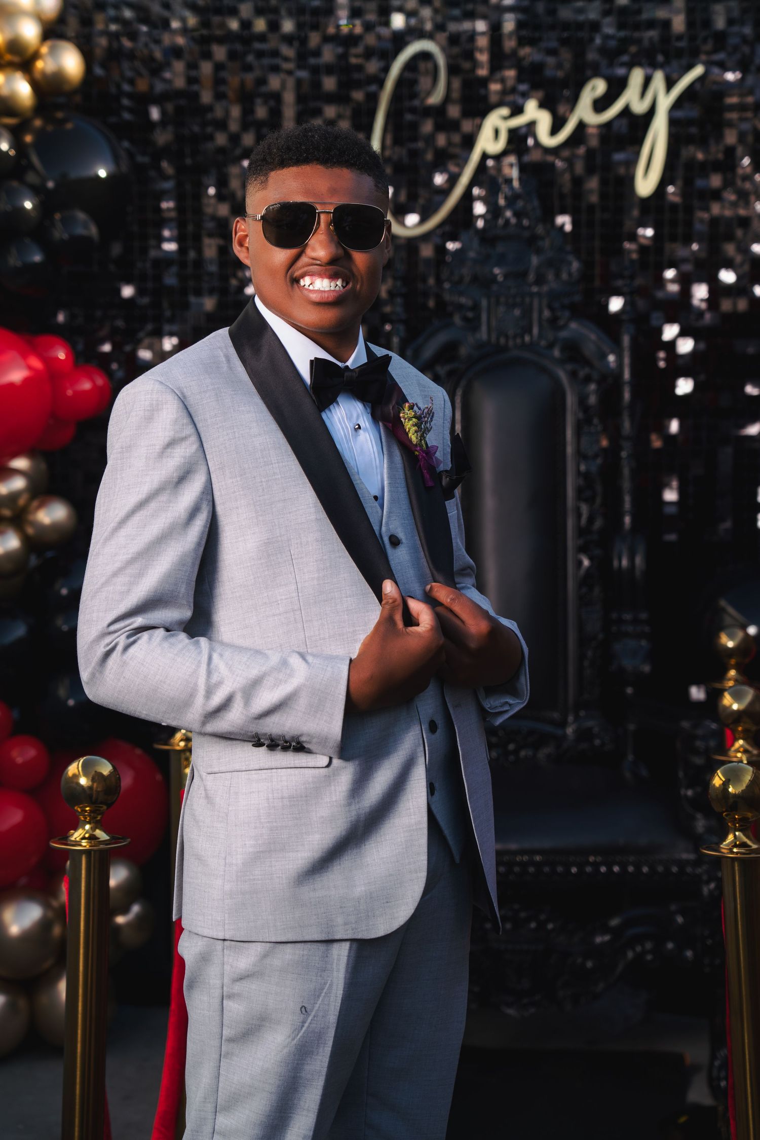 Young man in gray tuxedo with black lapels, bow tie, and sunglasses posing at prom event with balloon backdrop.