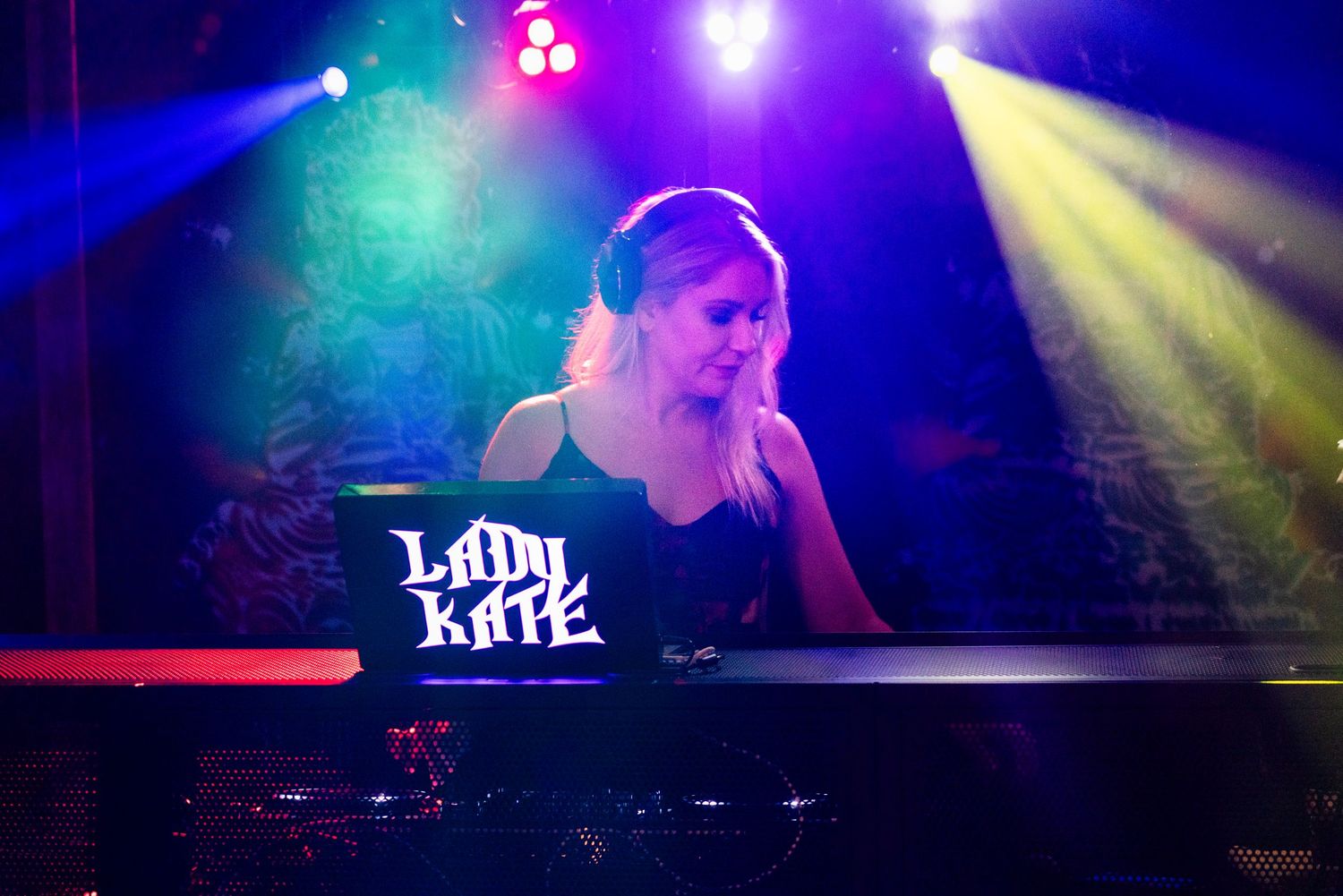 Female DJ Lady Kate performing at a nightclub under colorful stage lights wearing headphones.