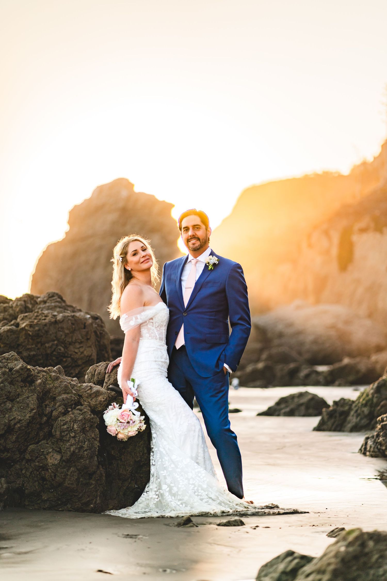 Couple in wedding attire embracing on rocky beach at sunset with golden light and ocean waves in background.