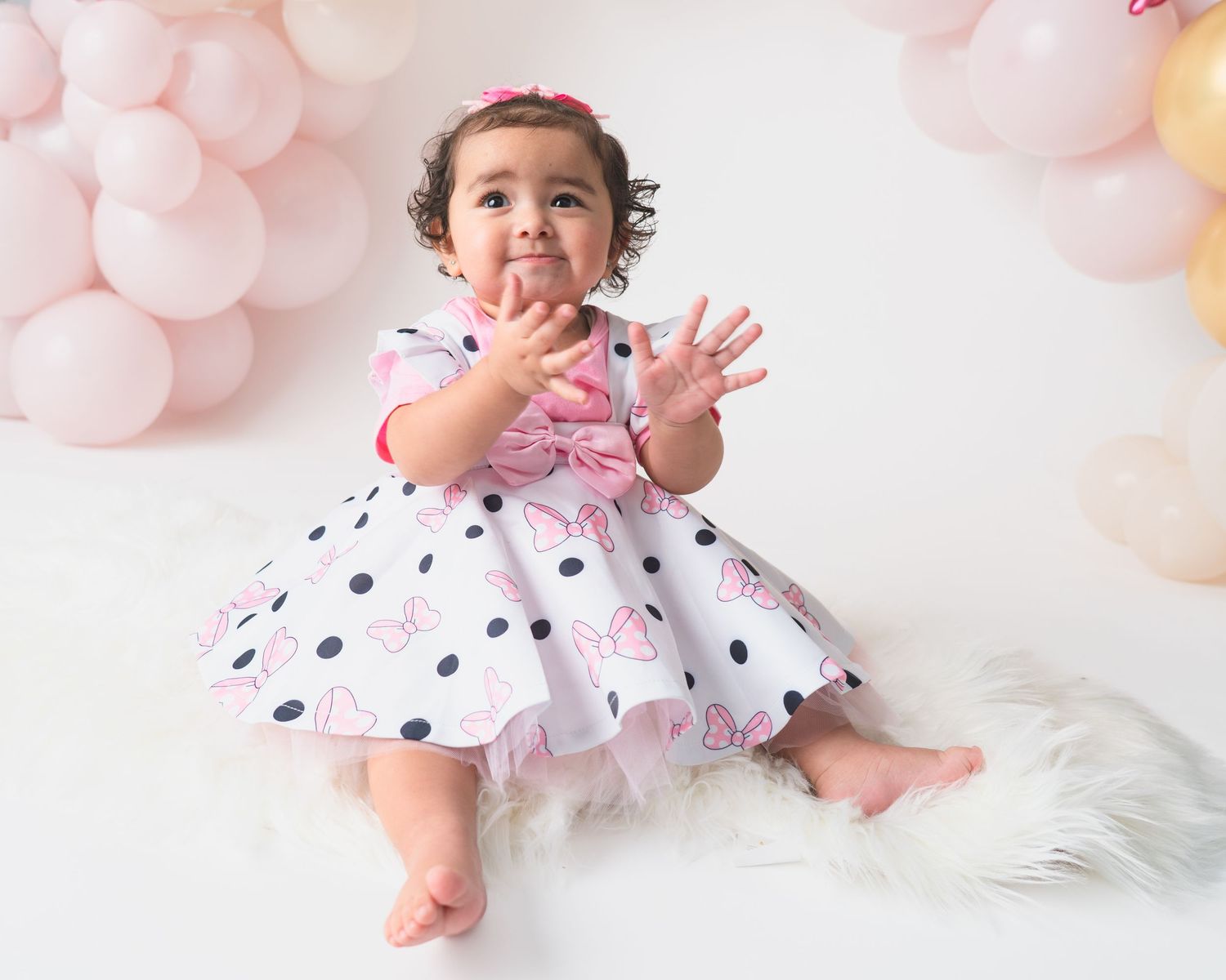 Baby in ruffled polka dot dress crawls on white floor with balloons.