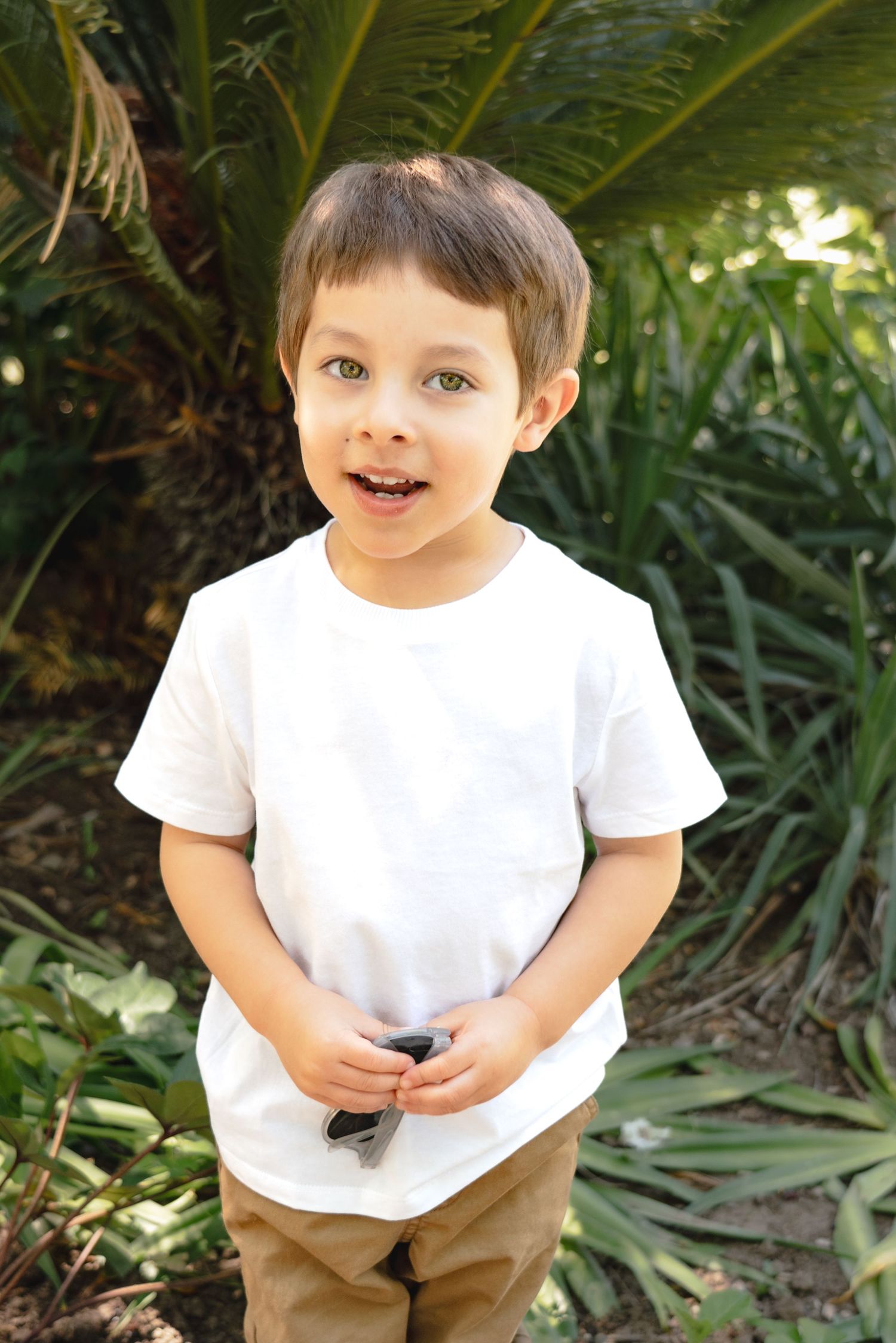 A child in a white t-shirt and khaki pants plays outdoors among tropical plants in a garden setting.