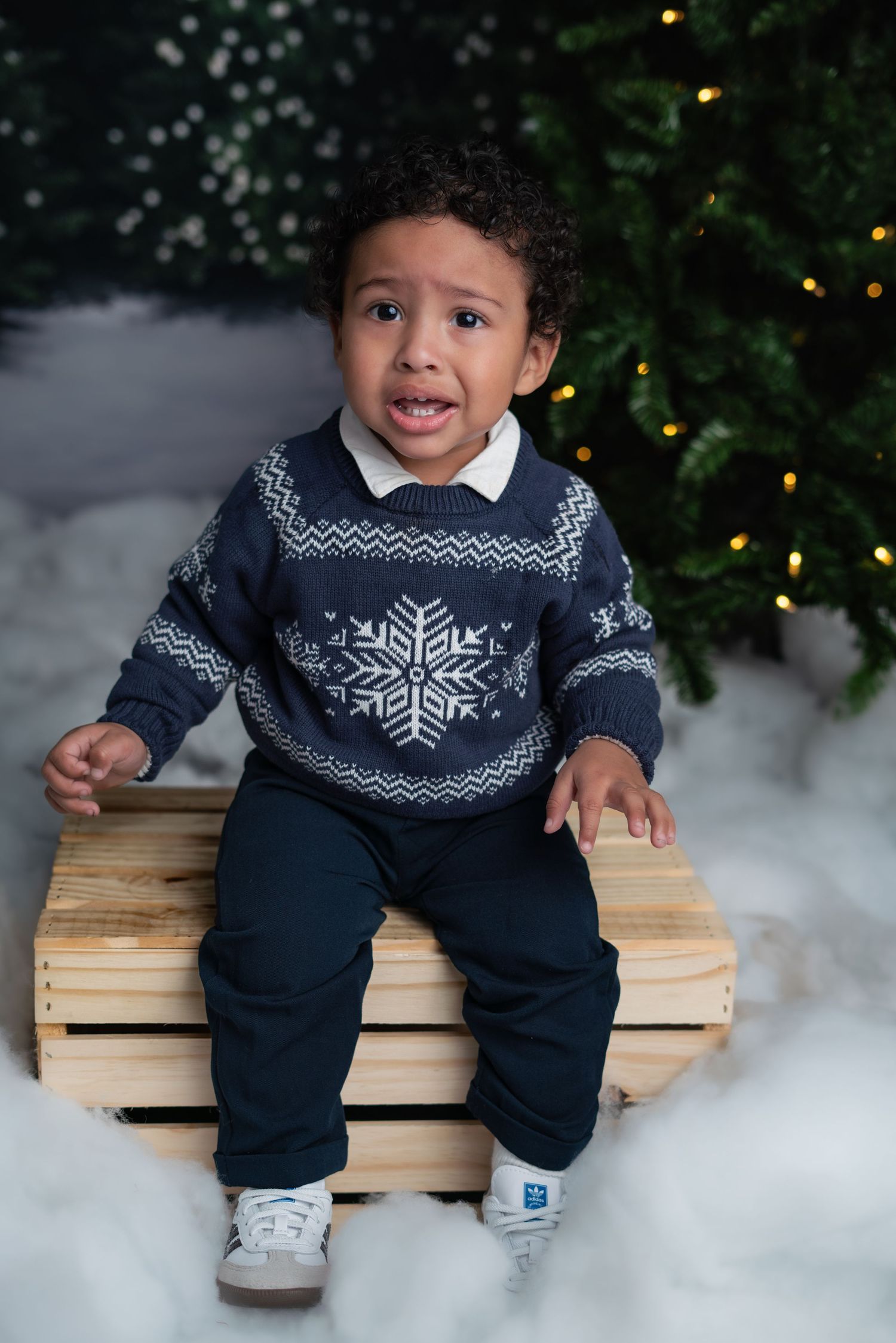 Child wearing festive navy snowflake sweater sits on wooden crate with Christmas tree and artificial snow in background.