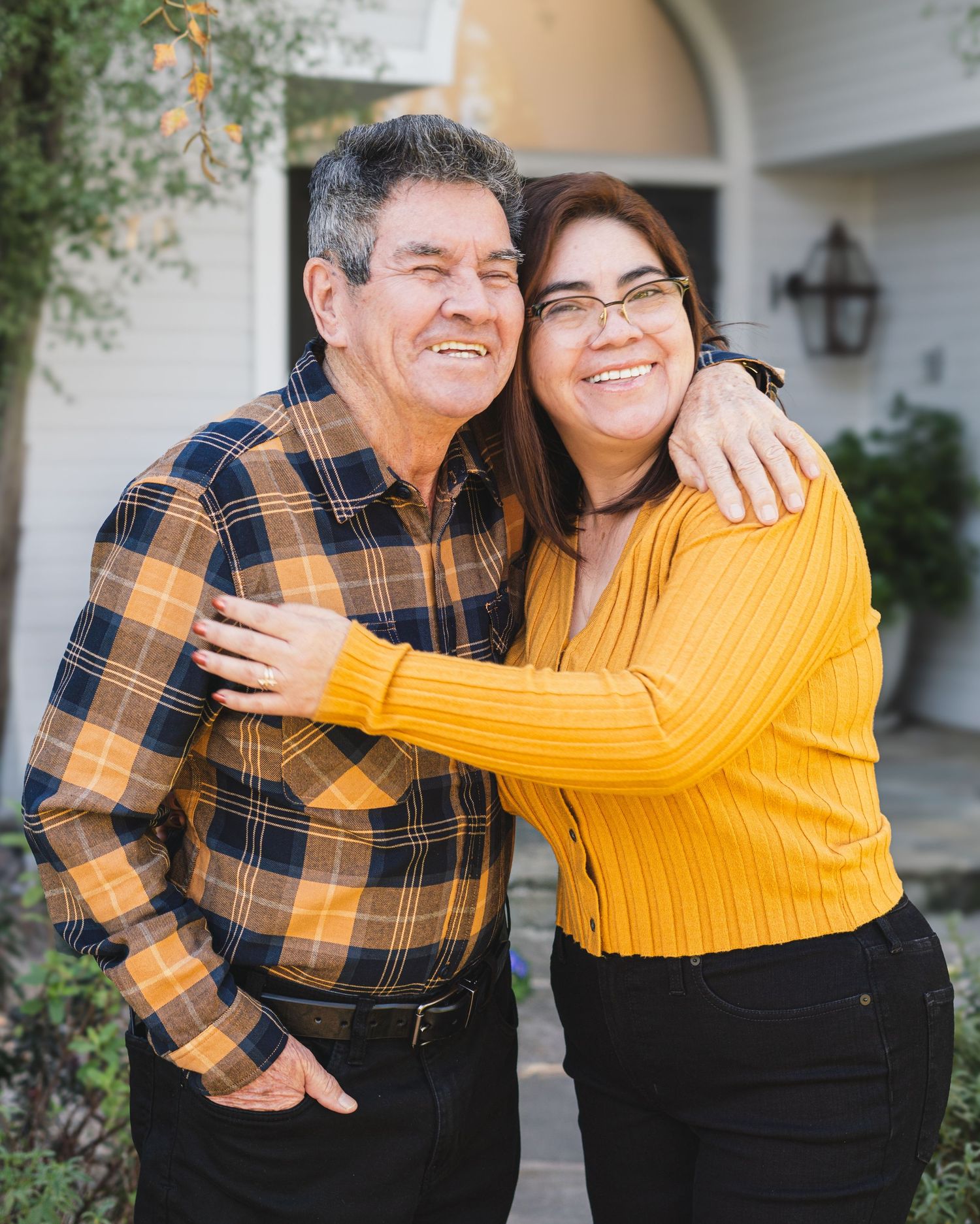 Two people sharing a warm embrace while wearing a yellow sweater and plaid shirt outdoors near a house.