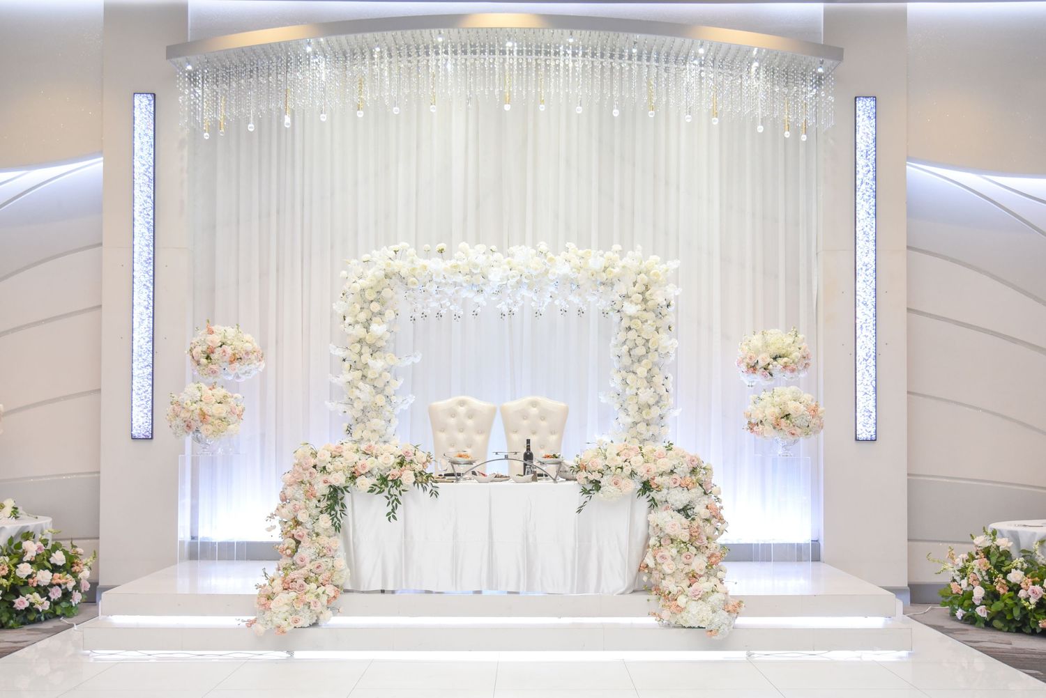 Luxurious white draped wedding ceremony backdrop with crystal chandeliers and floral arch decorations.