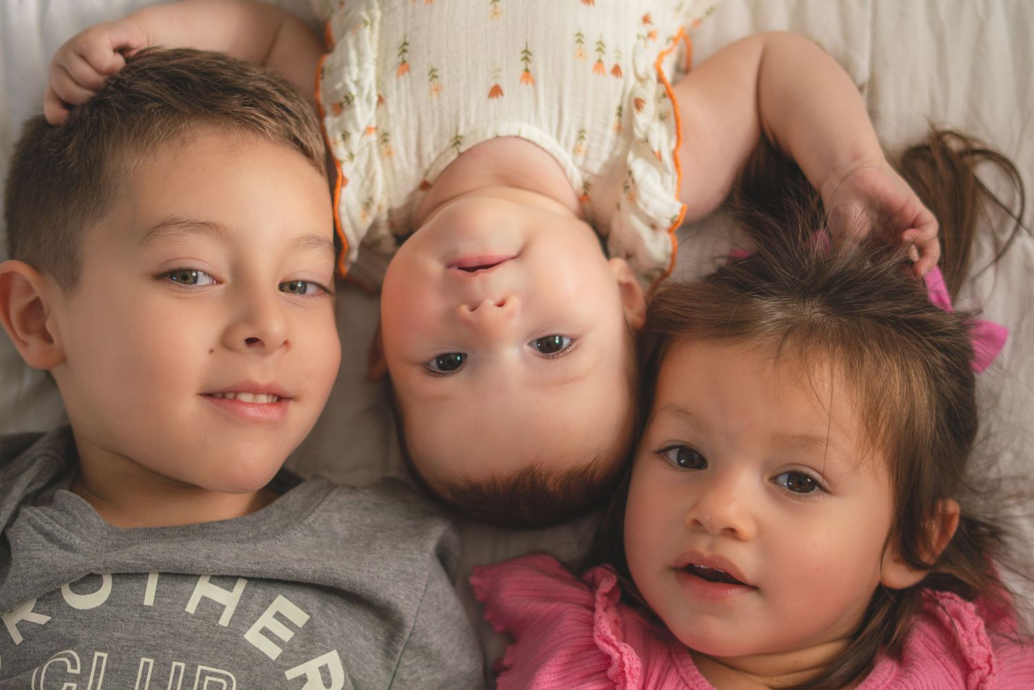 Three young siblings lying together on a white bed, smiling and looking up at the camera in a candid family portrait.