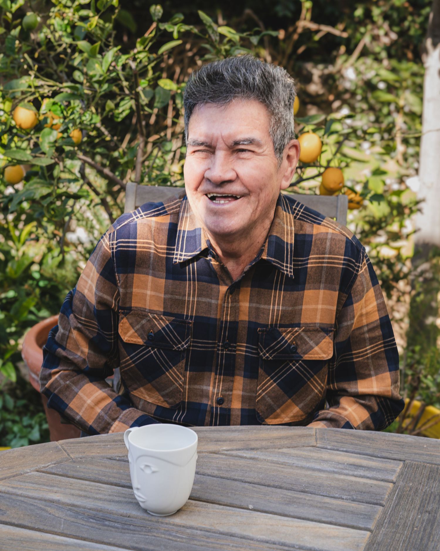 A person in a plaid flannel shirt sits at a wooden table in a garden with citrus trees.