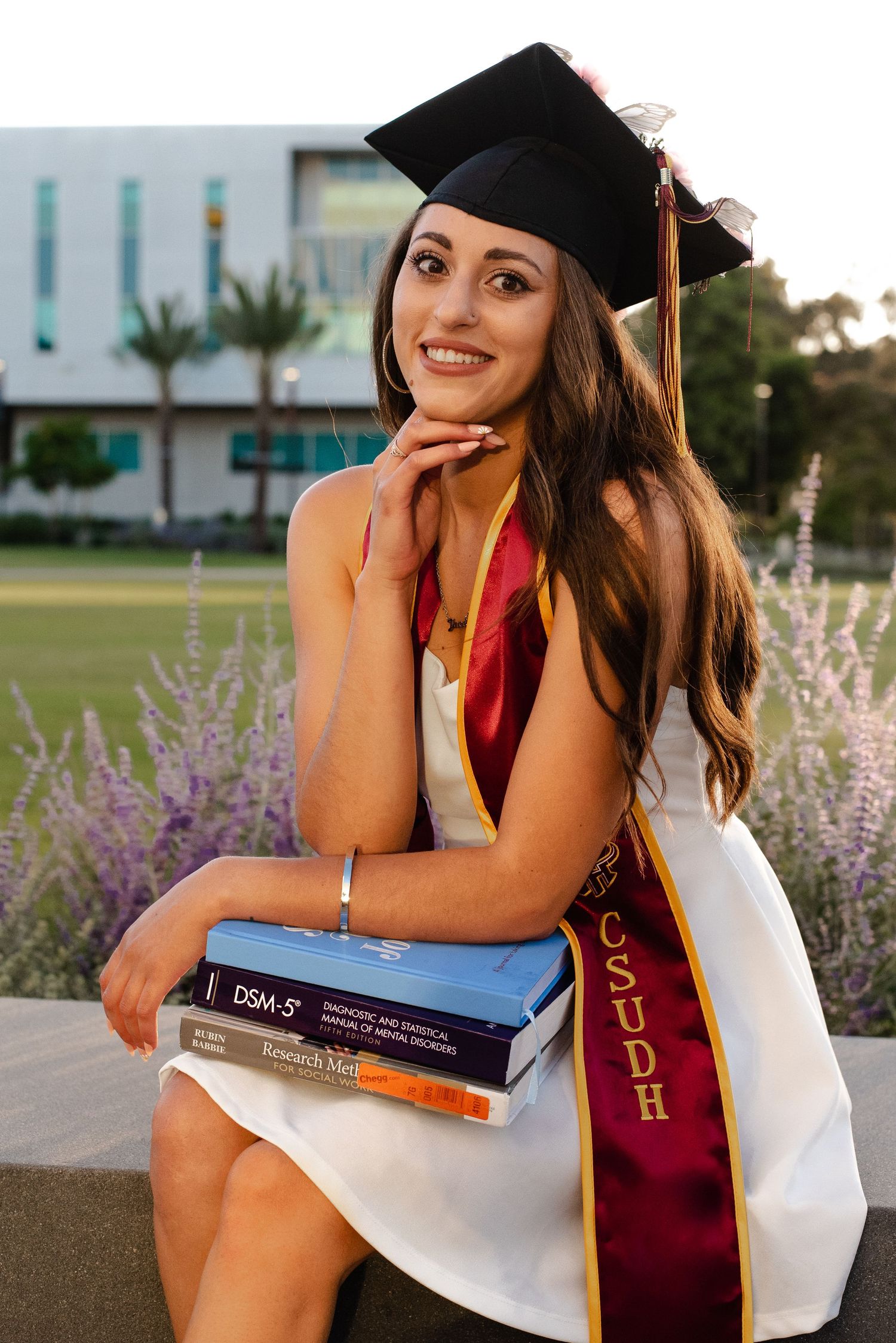 Graduate in cap and gown poses with textbooks on campus grounds.