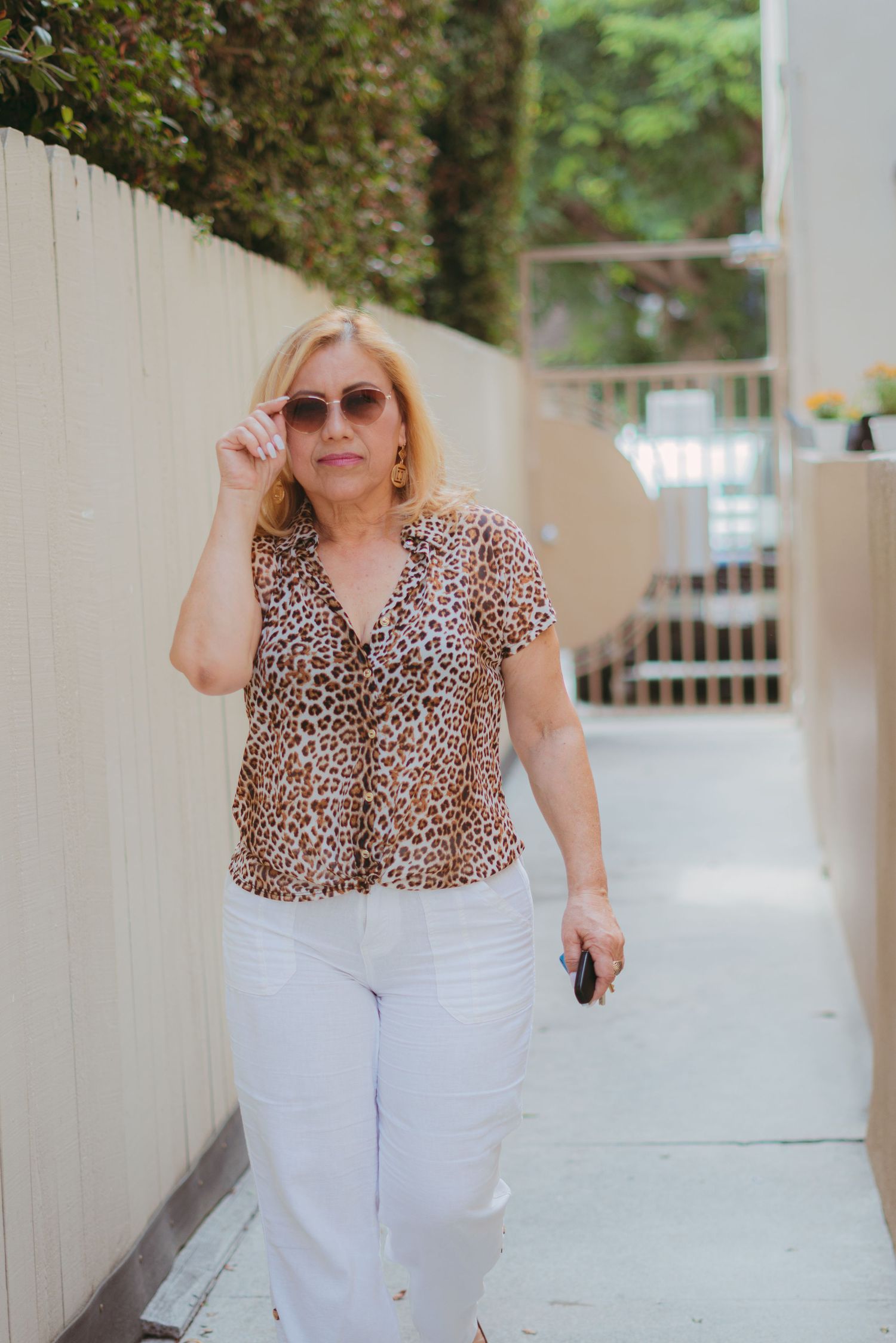 A person wearing a leopard print top and white pants poses against a light colored wall in an outdoor setting.