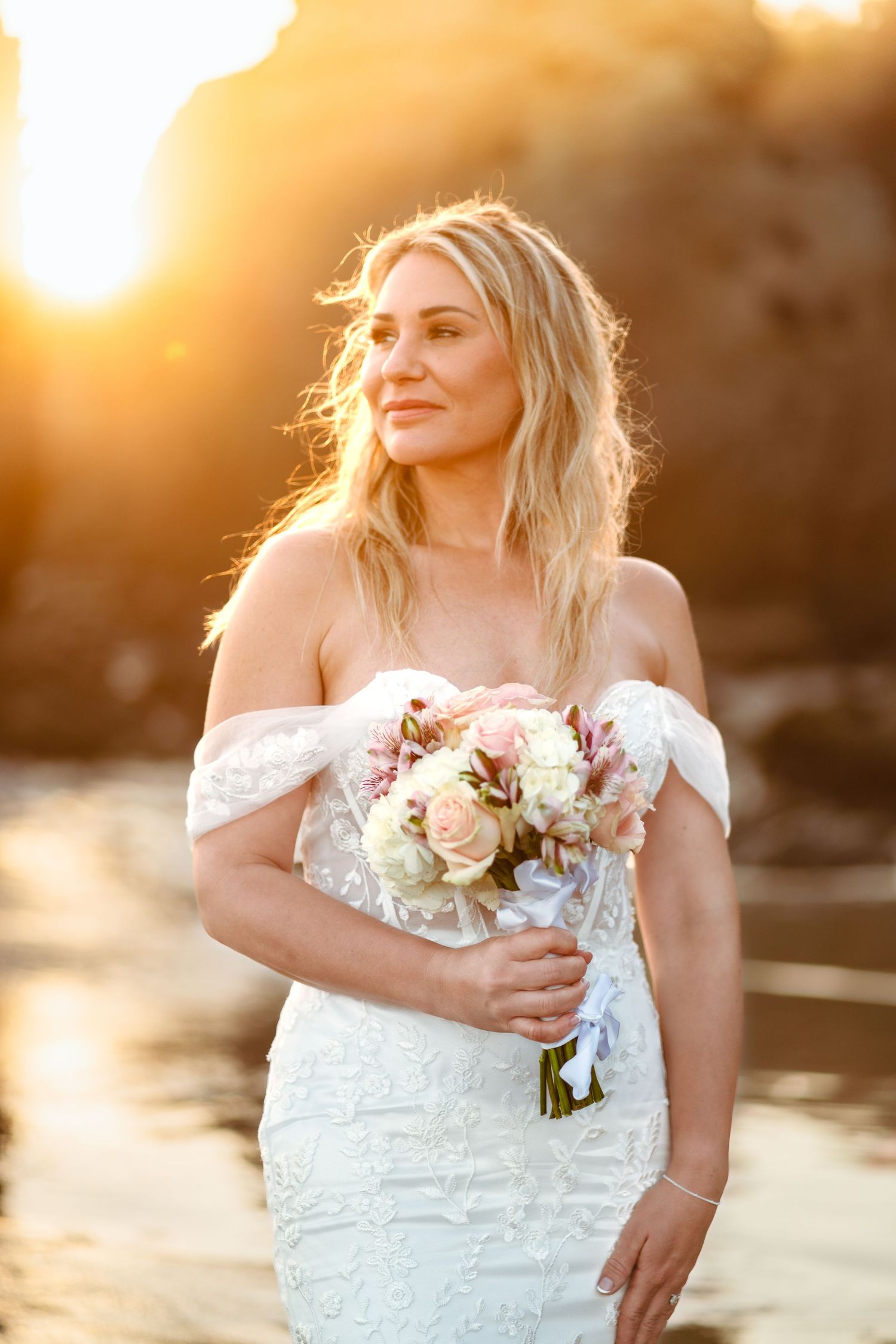 Bride in lace off-shoulder wedding dress holds romantic bouquet of roses during golden hour sunset photoshoot.