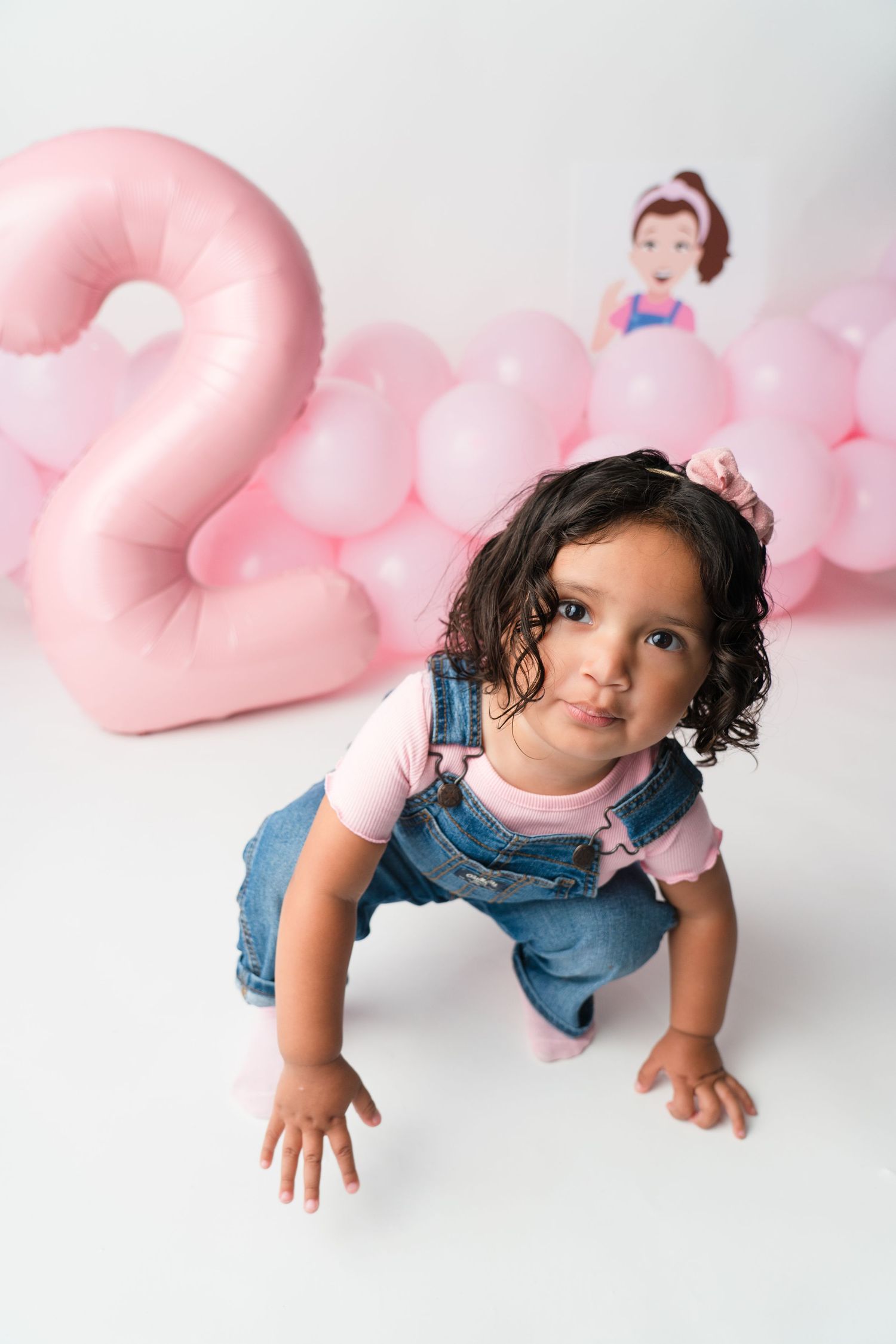 Child in denim crawling on white floor with pink heart balloons in the background.