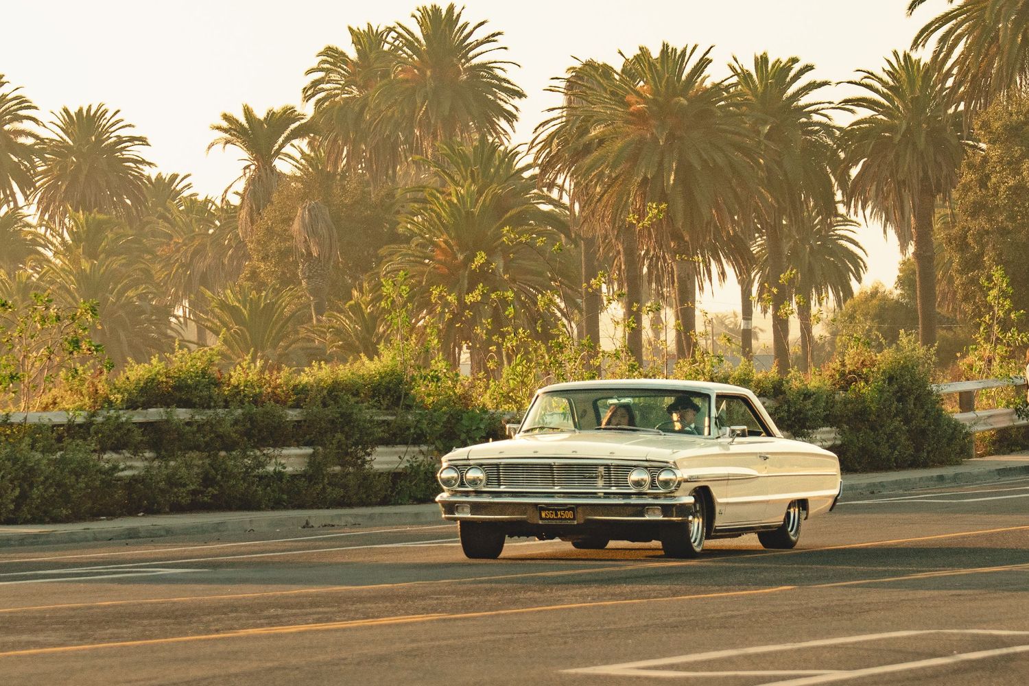 Vintage white classic car cruising past lush palm trees on a sunny California boulevard.