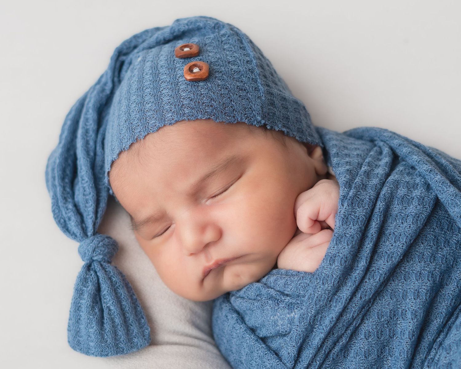 Sleeping newborn baby wrapped in a soft blue knit blanket and matching hat with wooden buttons.