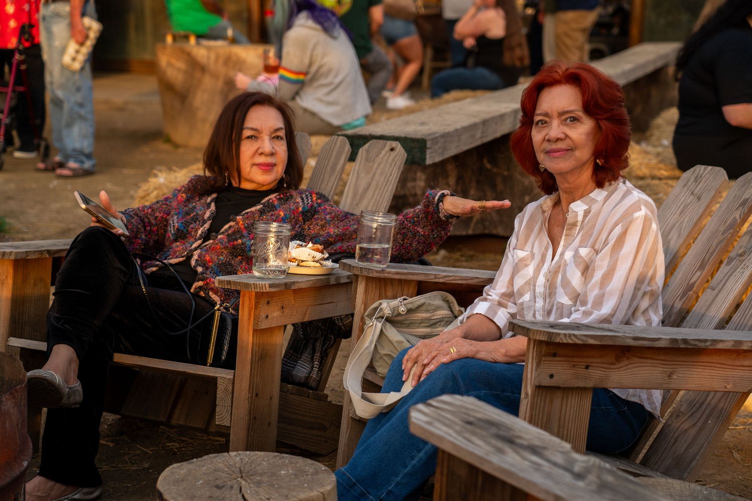 Two women smiling and relaxing at an outdoor wooden table during an evening social gathering.