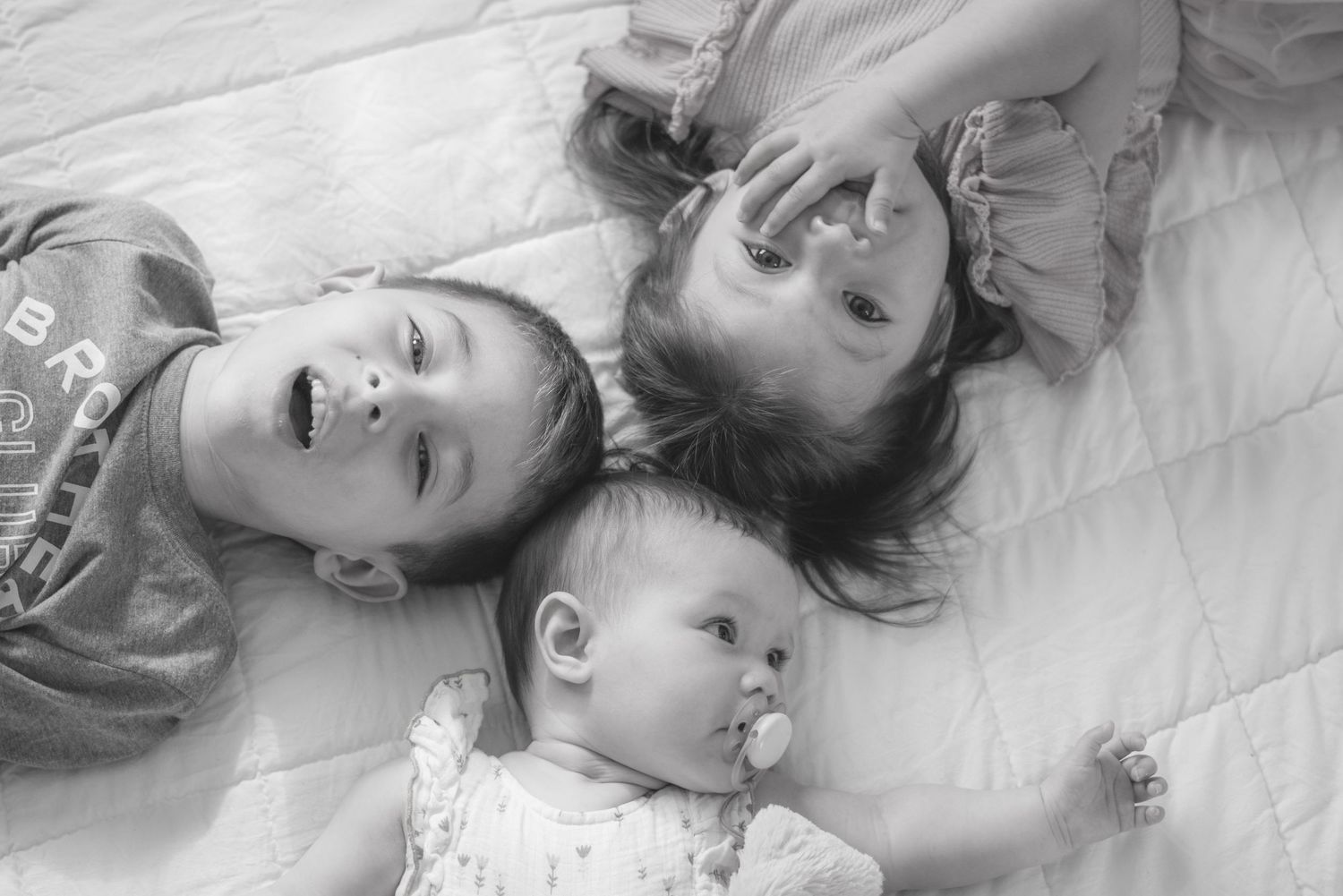 Black and white photo of three young children lying together on a blanket, two toddlers and a baby with a pacifier.