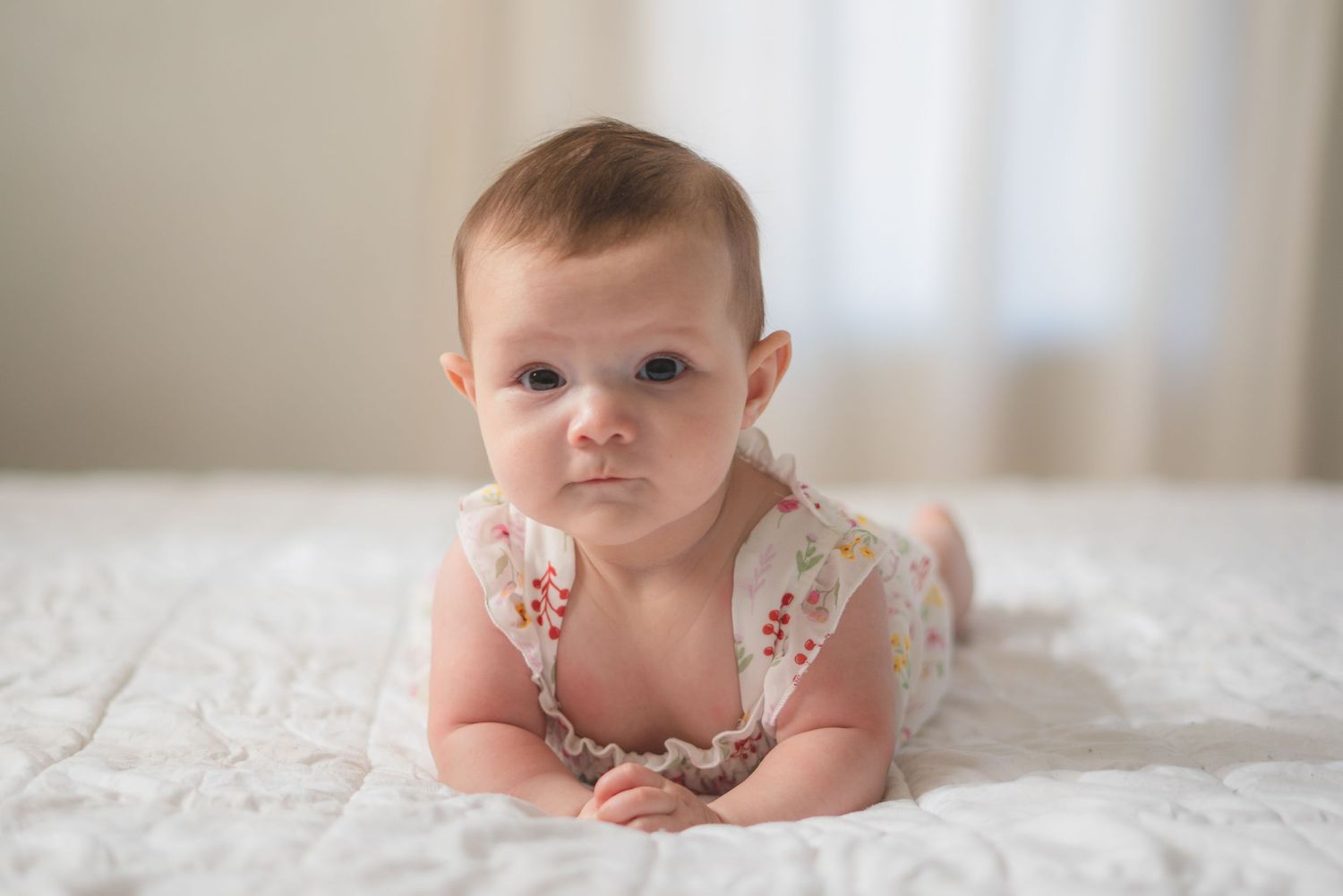 Cute baby girl lying on her tummy on a white bed, wearing a floral onesie, with a serious expression.