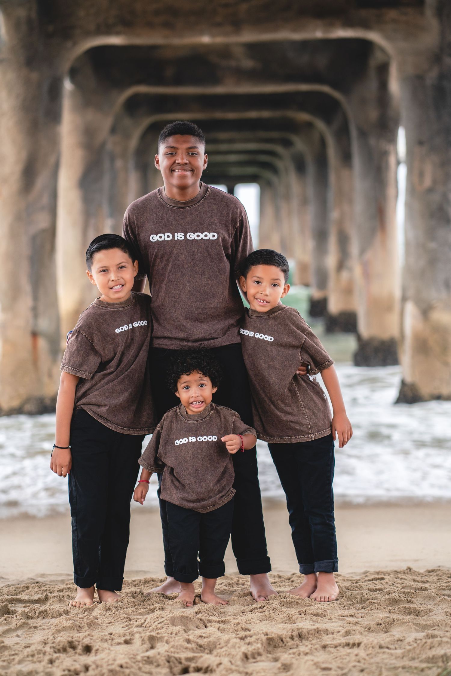 Four children in matching brown God Is Good shirts pose together on sandy beach under a pier.