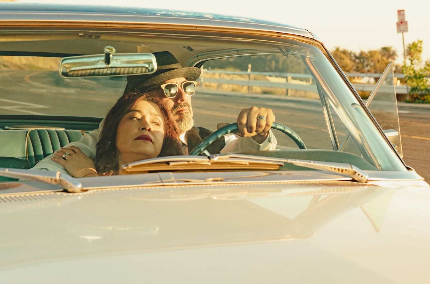 Two people in a classic convertible car, woman leaning on man wearing a hat, sunny outdoor setting.