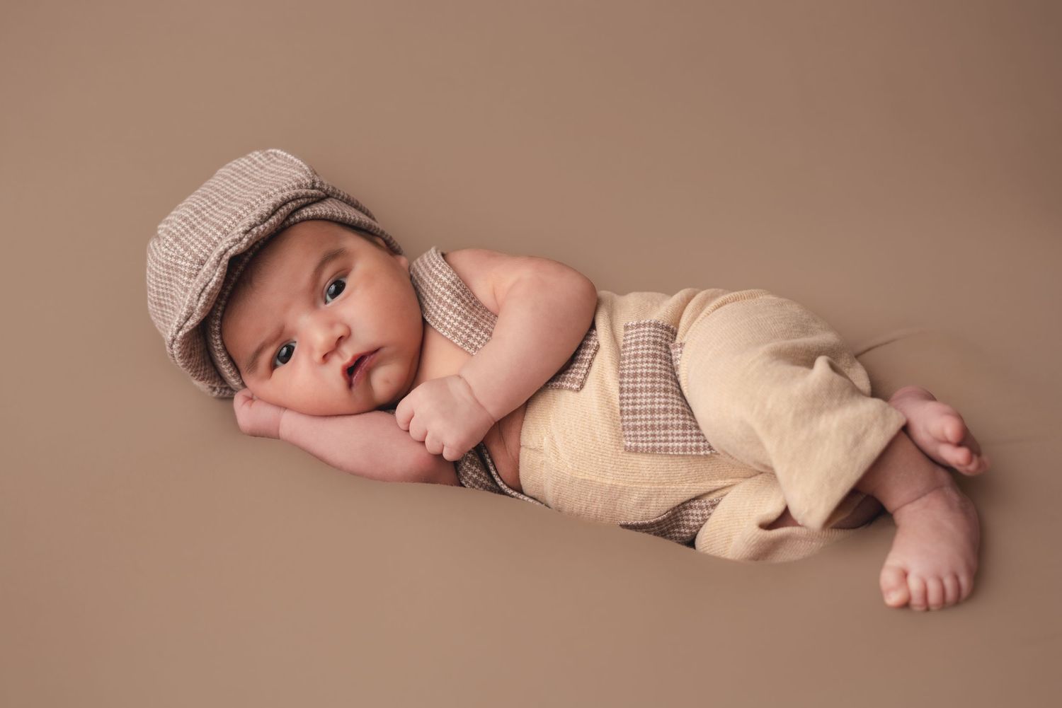Newborn baby in cream suspender outfit and plaid flat cap posed on brown backdrop.
