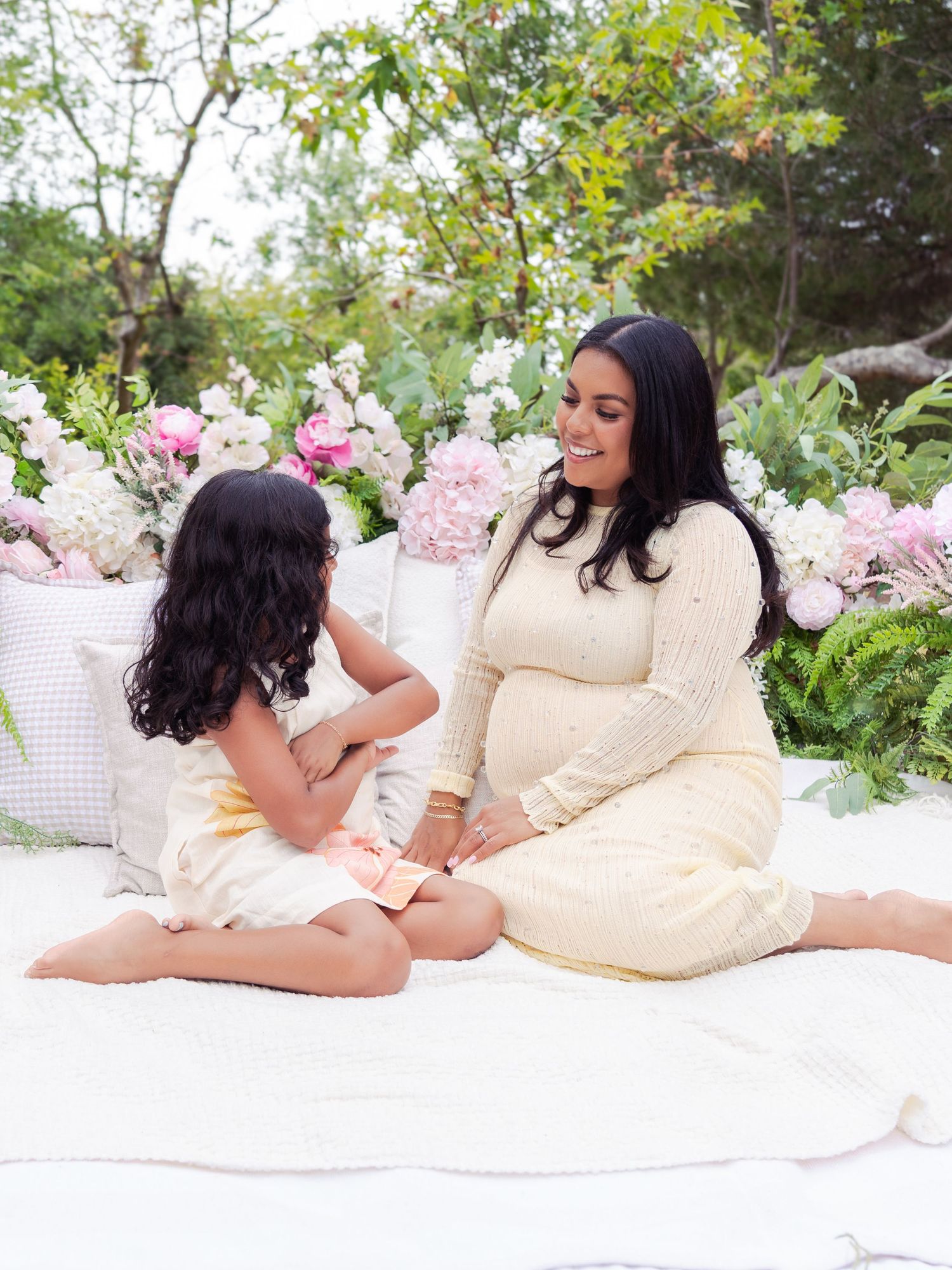 Person in cream dress relaxes on white blanket amid outdoor garden setting with pink floral accents.