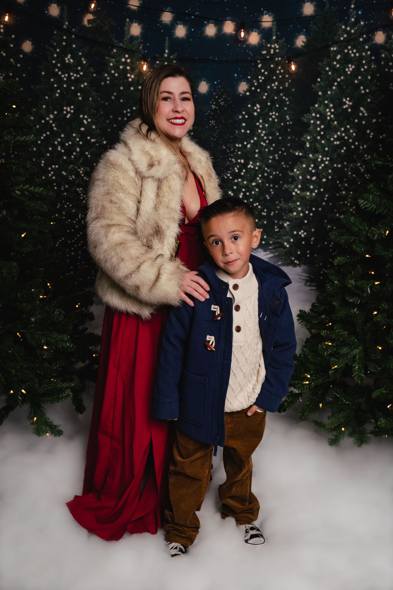 Holiday portrait scene with red dress, fur shawl, and navy suit against snowy backdrop with twinkling lights and evergreens.