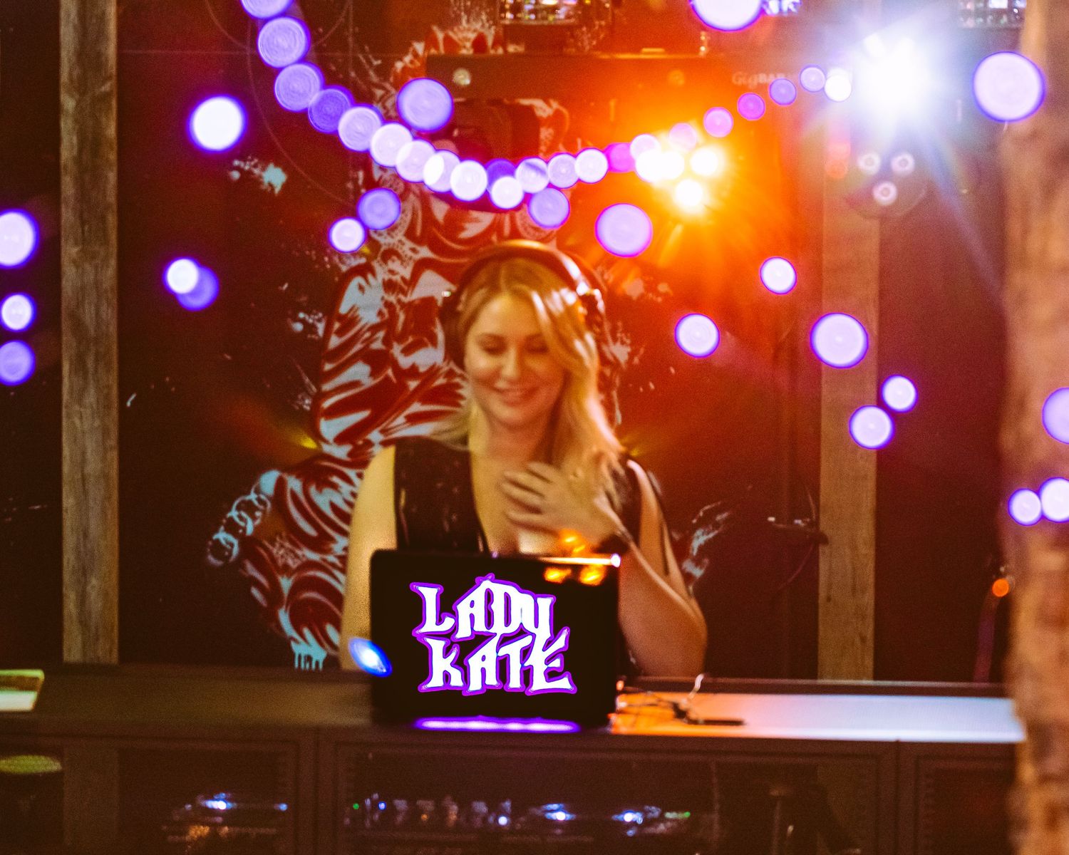 Female DJ Lady Kate smiling behind decks with headphones, colorful bokeh lights glowing in background.