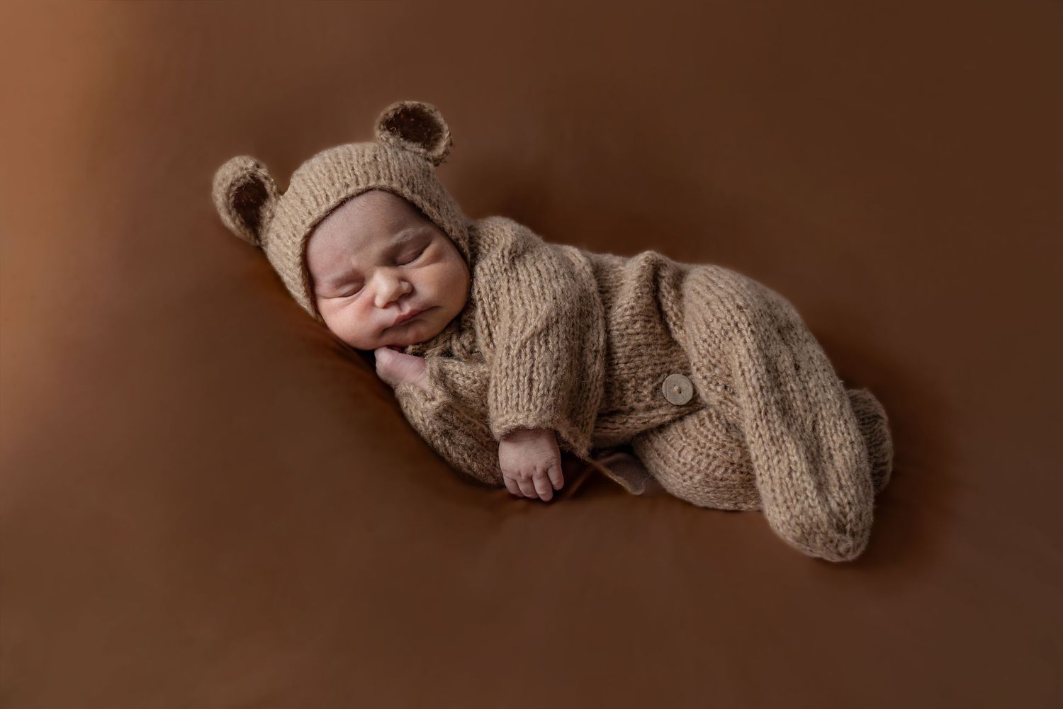 A newborn baby wearing a brown fuzzy teddy bear outfit lies peacefully on a soft brown background.