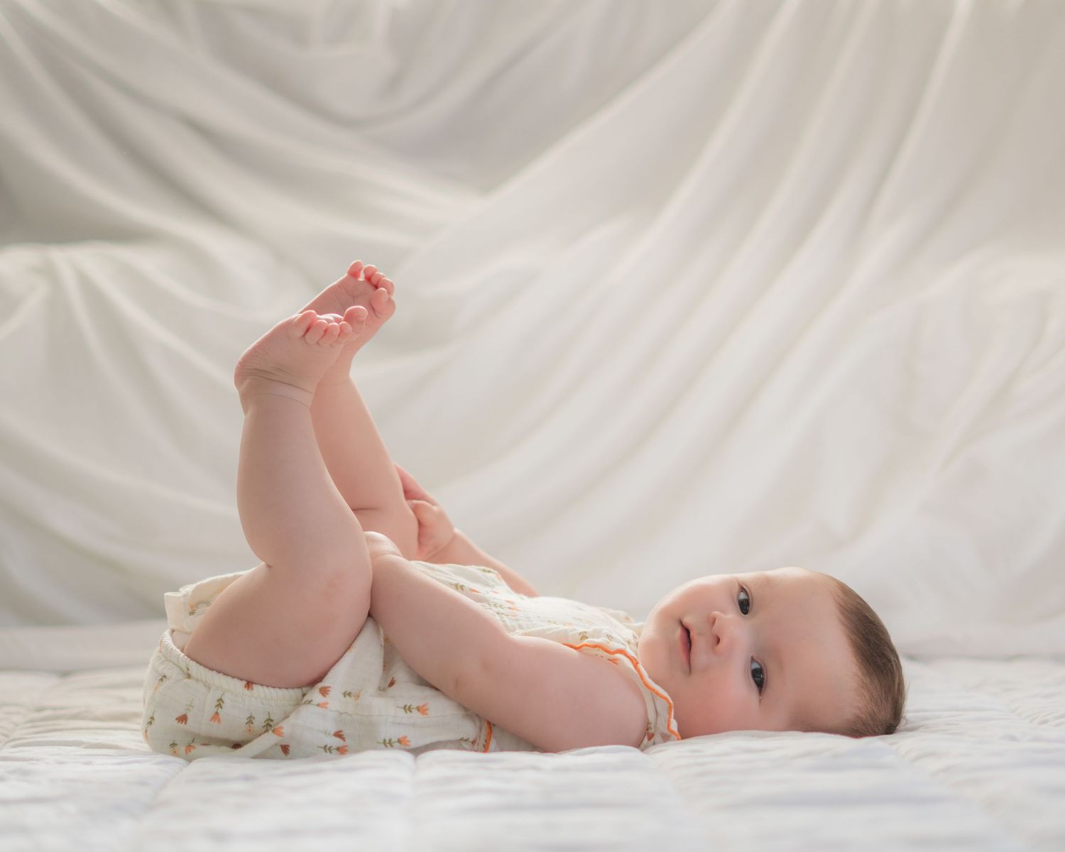 Adorable baby lying on back on white bedding, holding feet up, wearing floral diaper cover.