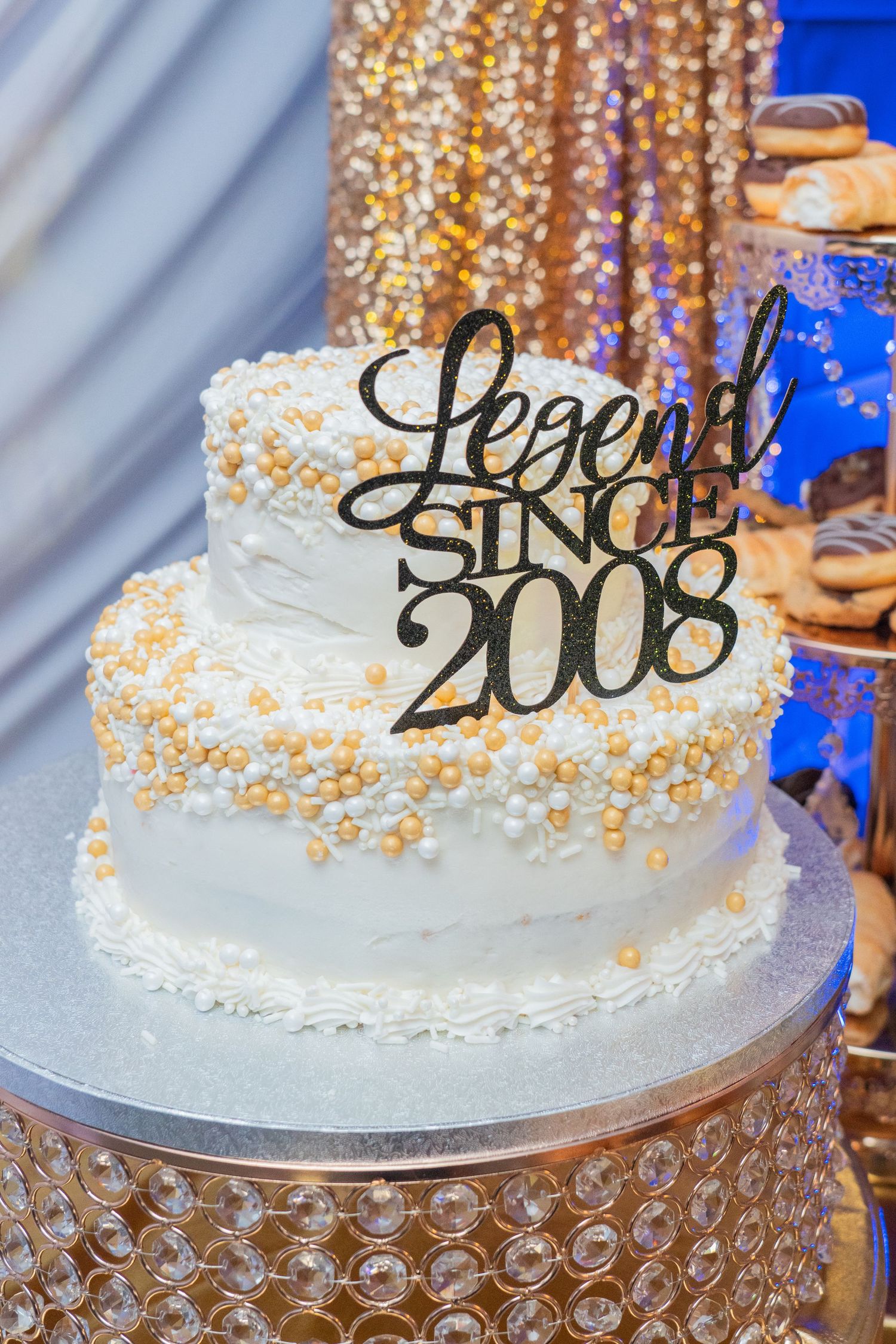 White celebration cake with gold pearls and black text decorations displayed on a crystal stand.