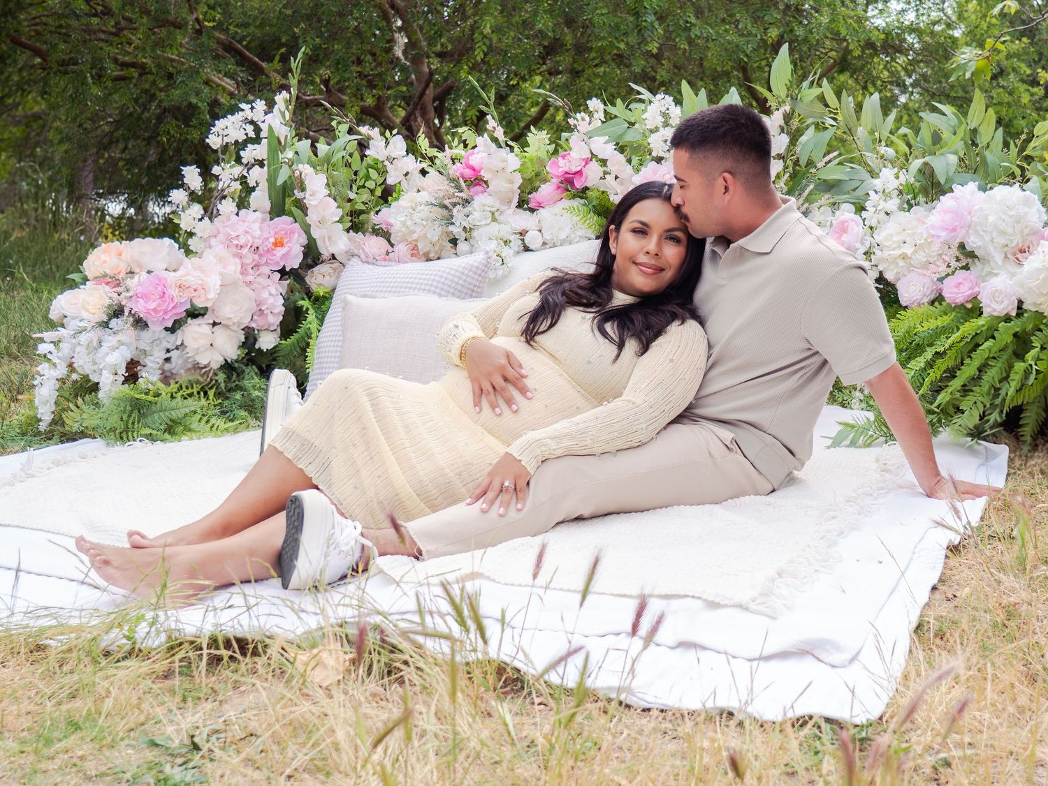 A romantic couple lounges on a white blanket surrounded by pink flowers in an outdoor setting.
