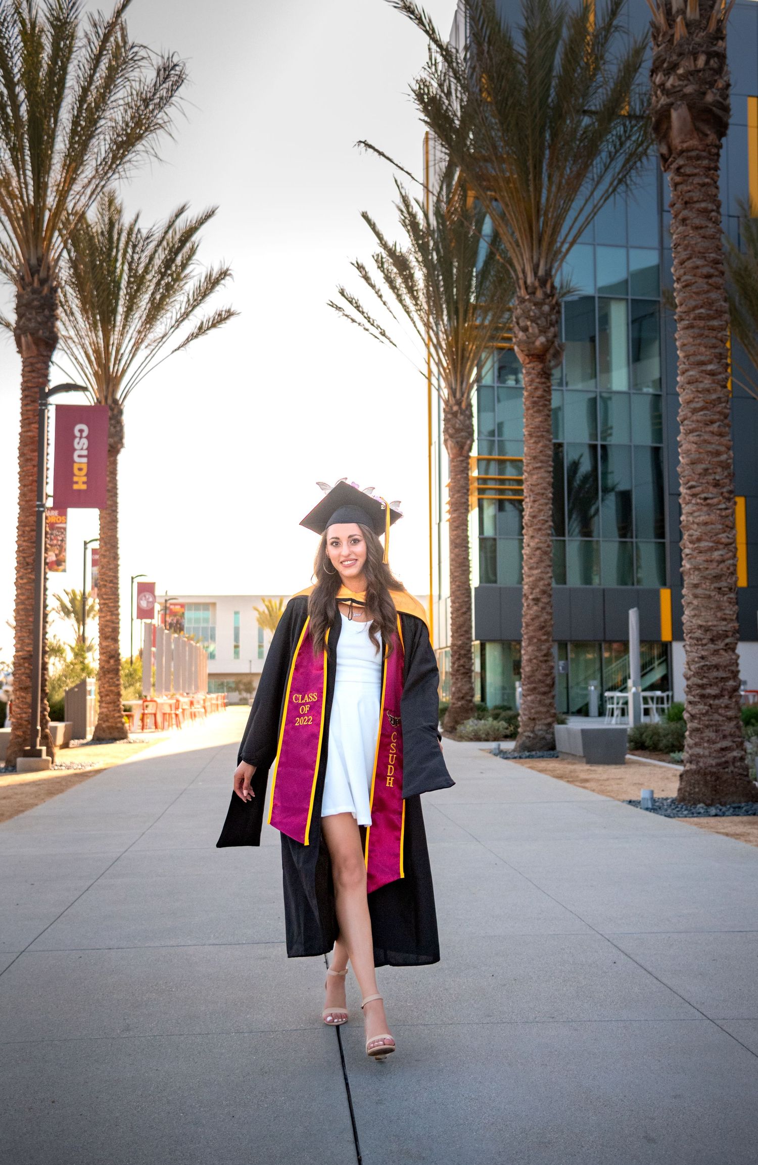 Graduate in academic regalia walks confidently on campus pathway lined with palm trees.