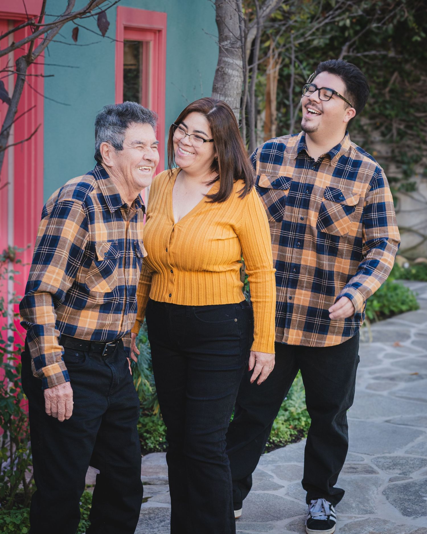 Three people wearing coordinating plaid shirts pose together against a turquoise building.