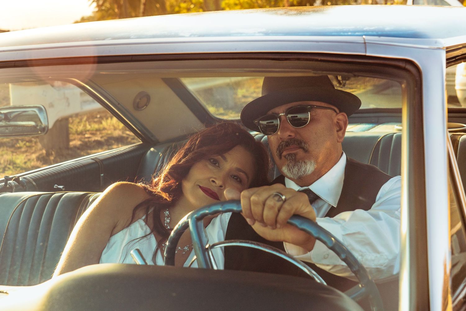 Couple in vintage car, man in hat and sunglasses with woman leaning on his shoulder, warm golden light.
