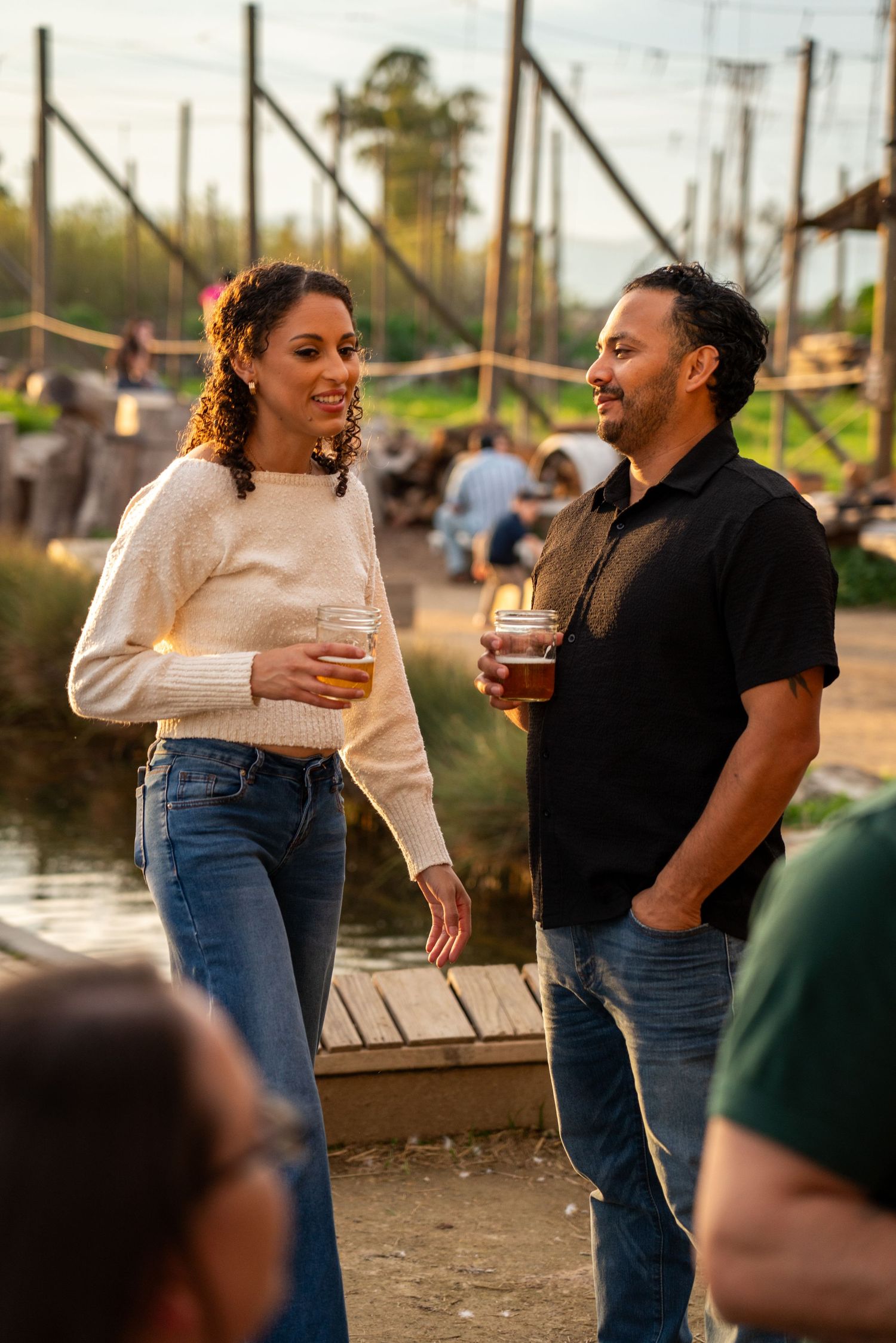 A man and woman socializing outdoors at a brewery, holding craft beers near a waterfront at golden hour.