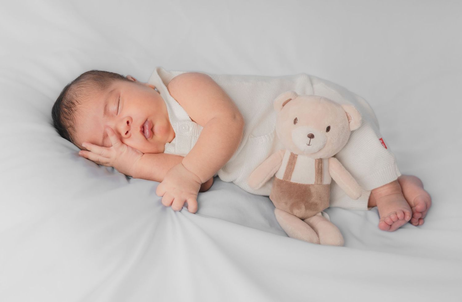Peaceful newborn baby sleeping beside a soft plush teddy bear toy on white background.