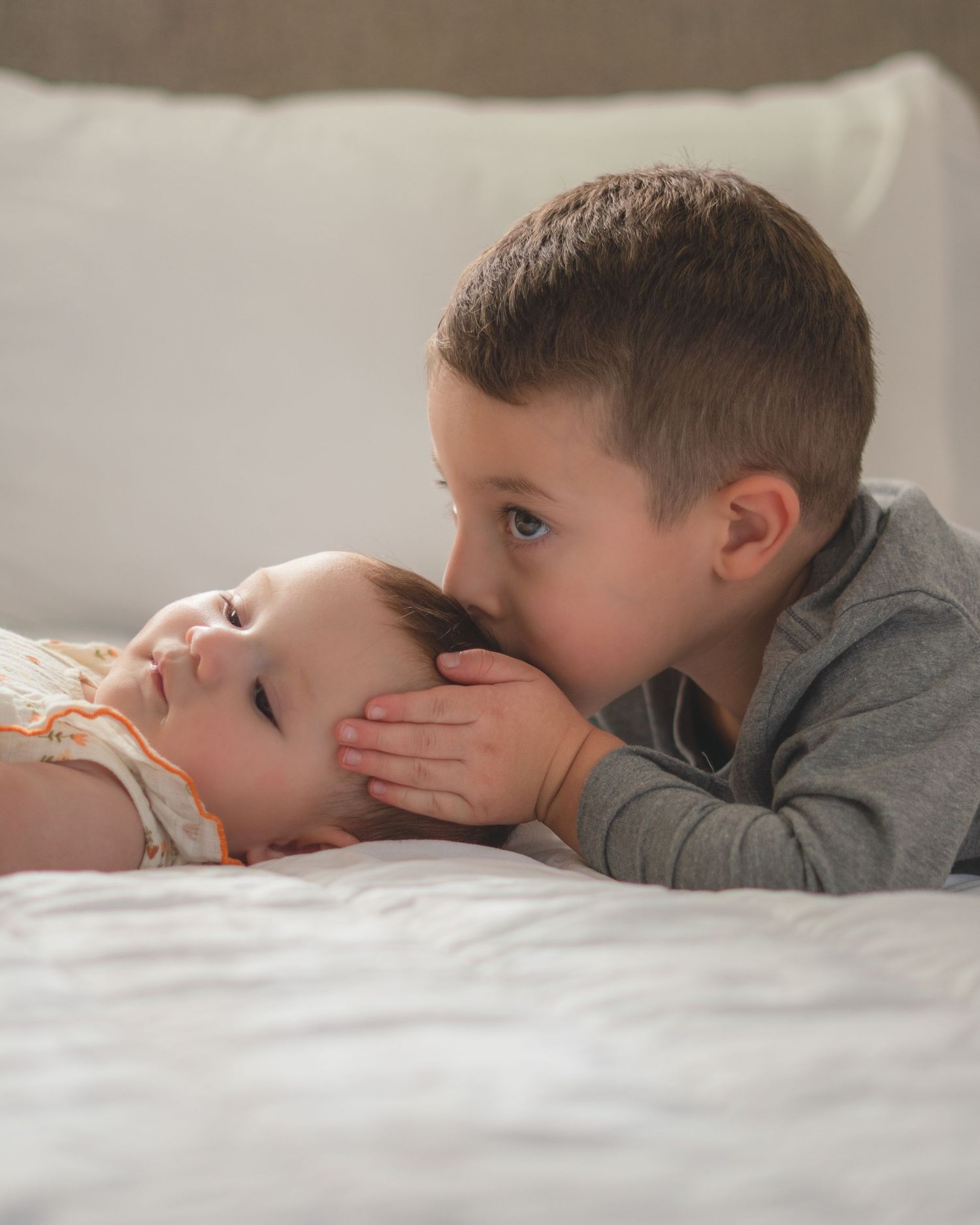 A young boy gently kisses a newborn baby on the head while lying on a white bed.
